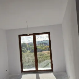 Wnętrze pokoju po świeżym pomalowaniu na biało, widok na zewnątrz przez drewniane okno z widocznym budynkiem Leroy Merlin w tle, surowy beton na podłodze i wystające przewody elektryczne.