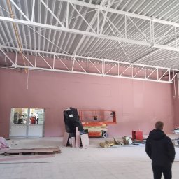 Marbud - Wnętrze hali w trakcie prac remontowych, widoczna podnośnik koszowy, materiały budowlane i osoba w oddali przy wejściu.