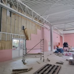 Marbud - Wnętrze hali przemysłowej w trakcie prac: ściany z izolacją z wełny mineralnej i różowych płyt, rusztowanie, paleta na podłodze, widoczne elementy konstrukcji dachu.