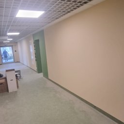 Marbud - Korytarz biurowy z pomalowanymi ścianami na beżowo, zielonymi wnękami drzwiowymi, podwieszanym sufitem kasetonowym i widocznymi elementami wyposażenia w oddali.