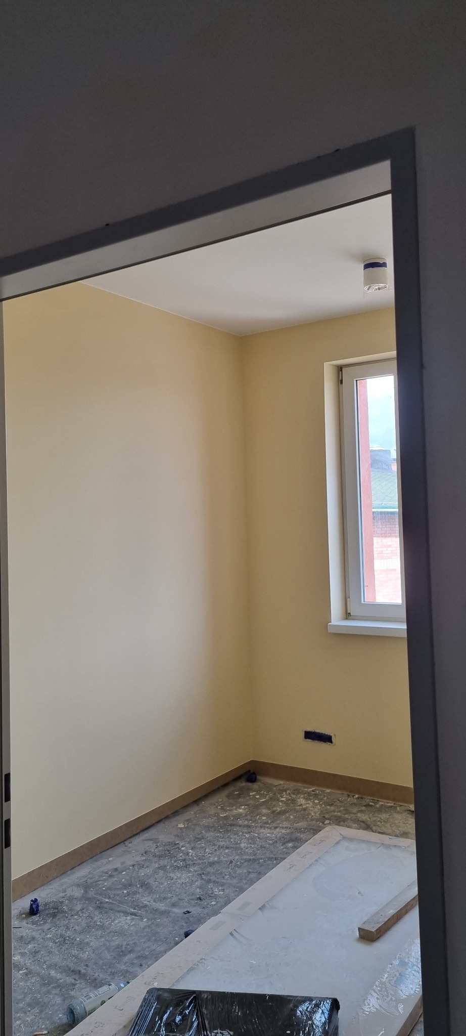 Pomalowane na jasnożółto ściany pokoju z oknem, widok zza framugi drzwi, podłoga przygotowana do dalszych prac wykończeniowych, widoczna kratka wentylacyjna w ścianie.