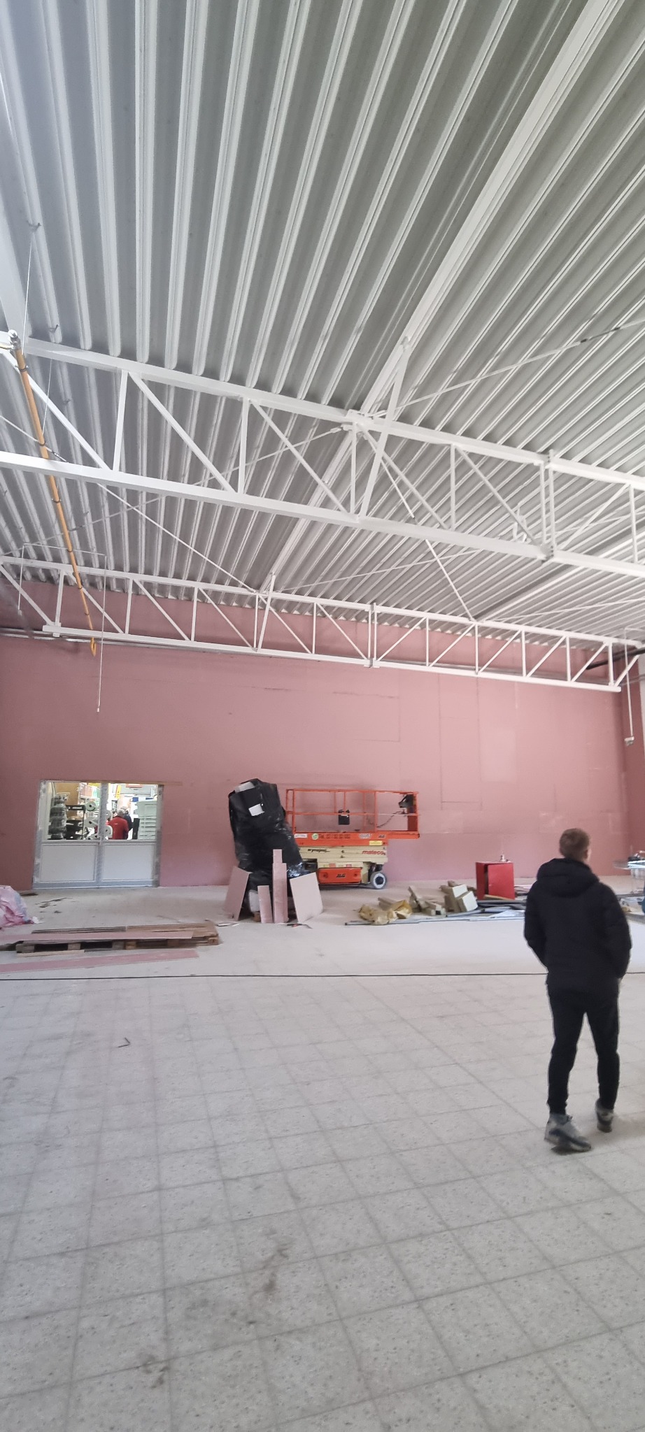 Wnętrze hali w trakcie prac remontowych, widoczna podnośnik koszowy, materiały budowlane i osoba w oddali przy wejściu.