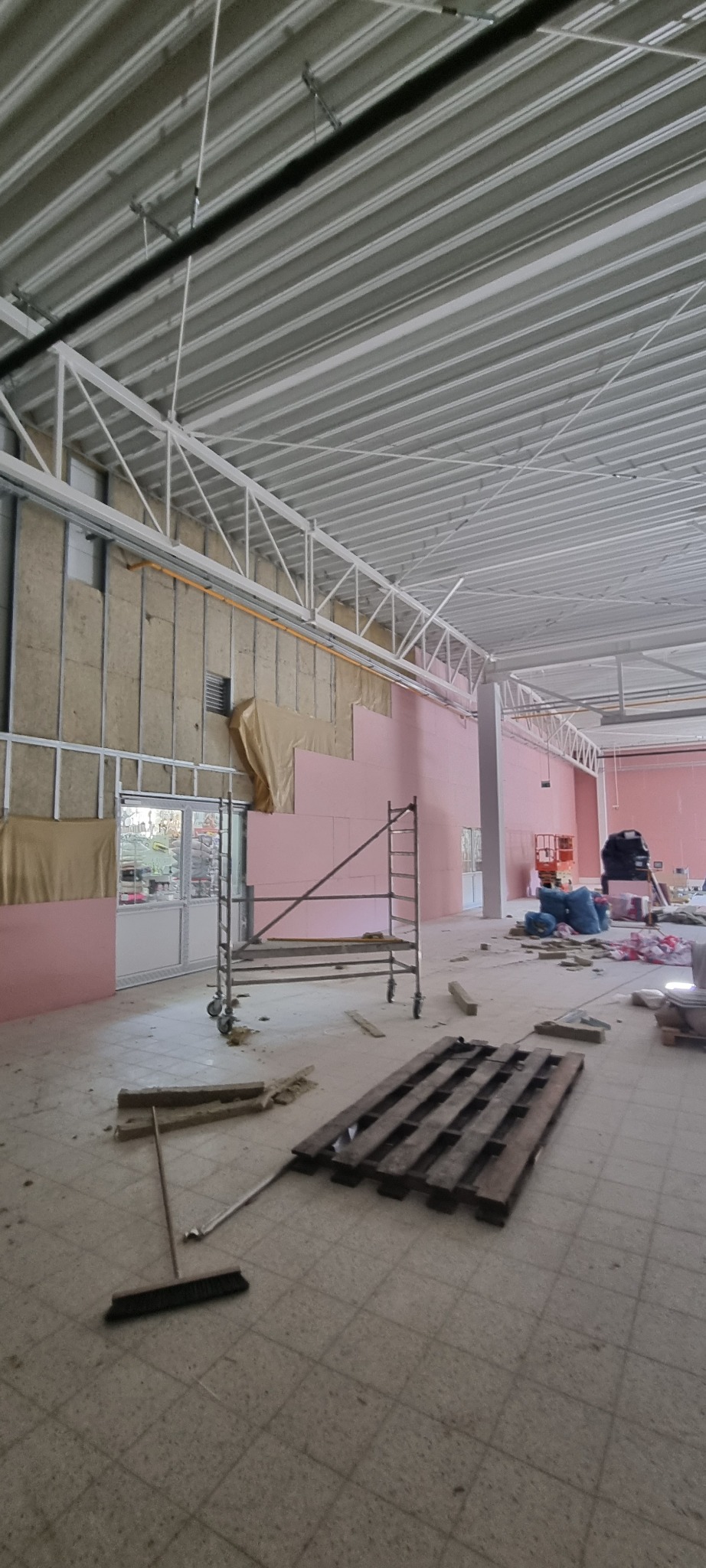 Wnętrze hali przemysłowej w trakcie prac: ściany z izolacją z wełny mineralnej i różowych płyt, rusztowanie, paleta na podłodze, widoczne elementy konstrukcji dachu.