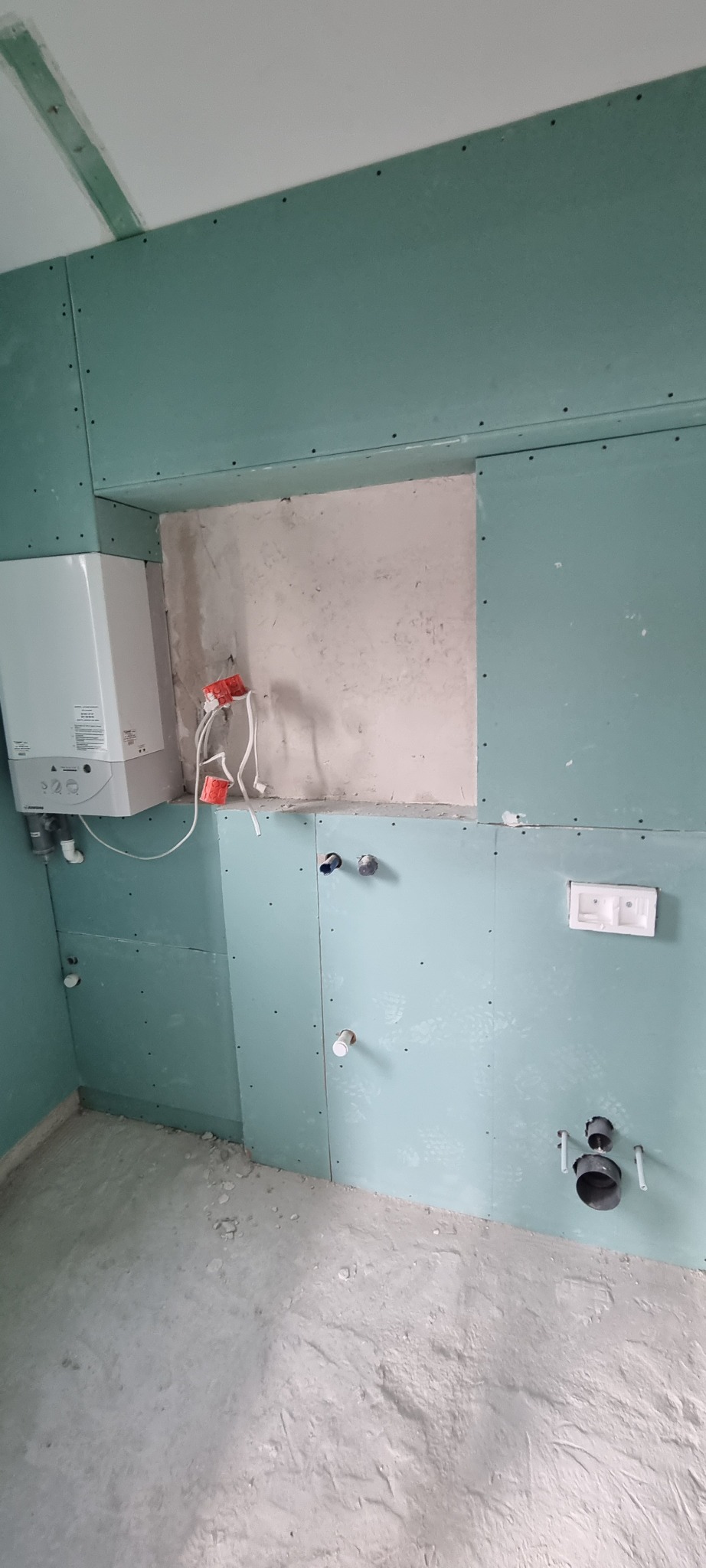 Wnętrze w trakcie wykańczania: ściany pokryte płytami gipsowo-kartonowymi w kolorze zielonym, widoczne instalacje hydrauliczne i elektryczne, fragment ściany z surowym tynkiem, podłoga wylewka.
