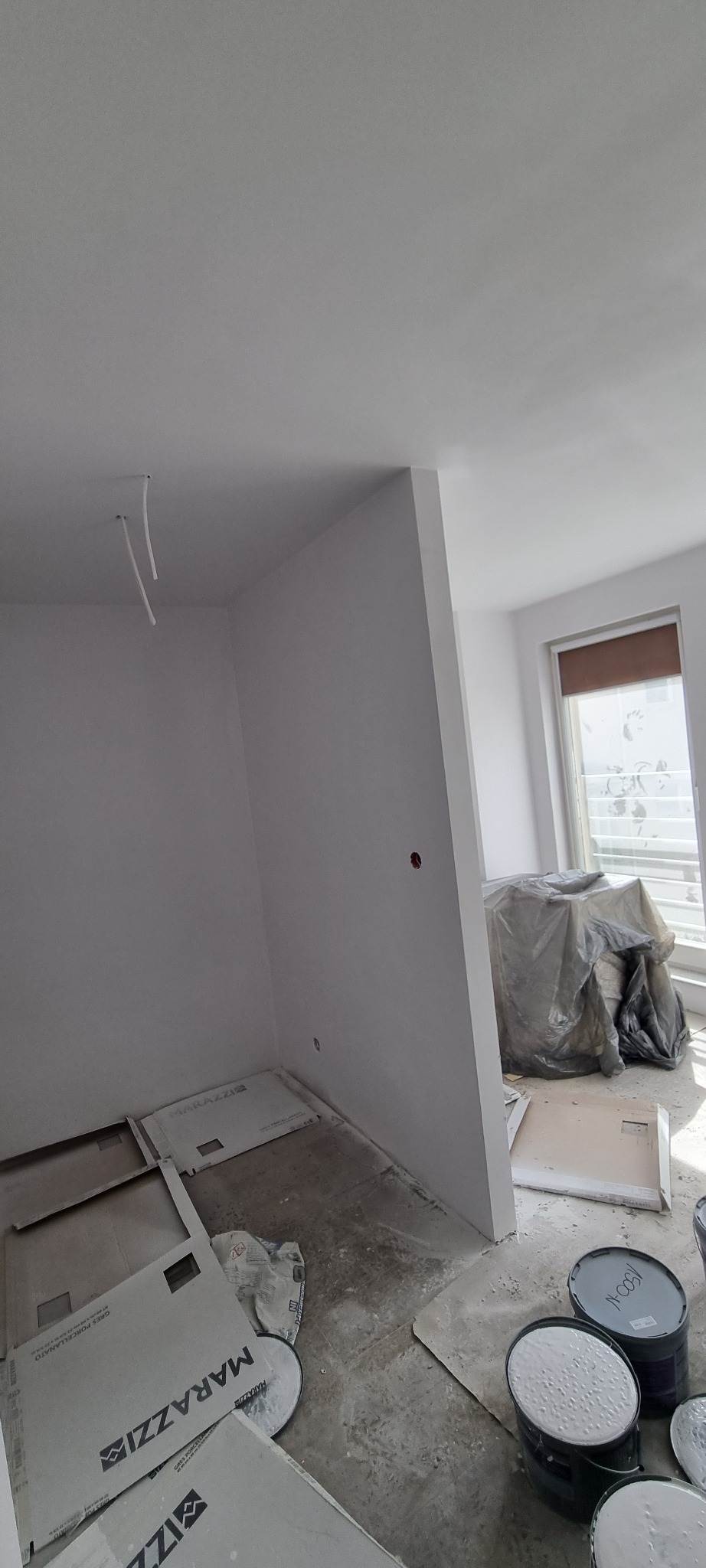 Wnętrze w trakcie prac wykończeniowych: świeżo pomalowane na biało ściany, zabezpieczone meble pod folią, puszki z farbą i płyty podłogowe na podłodze.