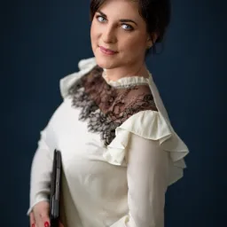 Portret kobiety w białej bluzce z koronkowym detalem, trzymającej tablet na tle ciemnoniebieskiej ściany.