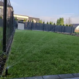 Automatyczny system nawadniania w akcji, zraszacz nawadnia świeżo założony trawnik z rolki, widoczny czarny płot i budynki w tle.