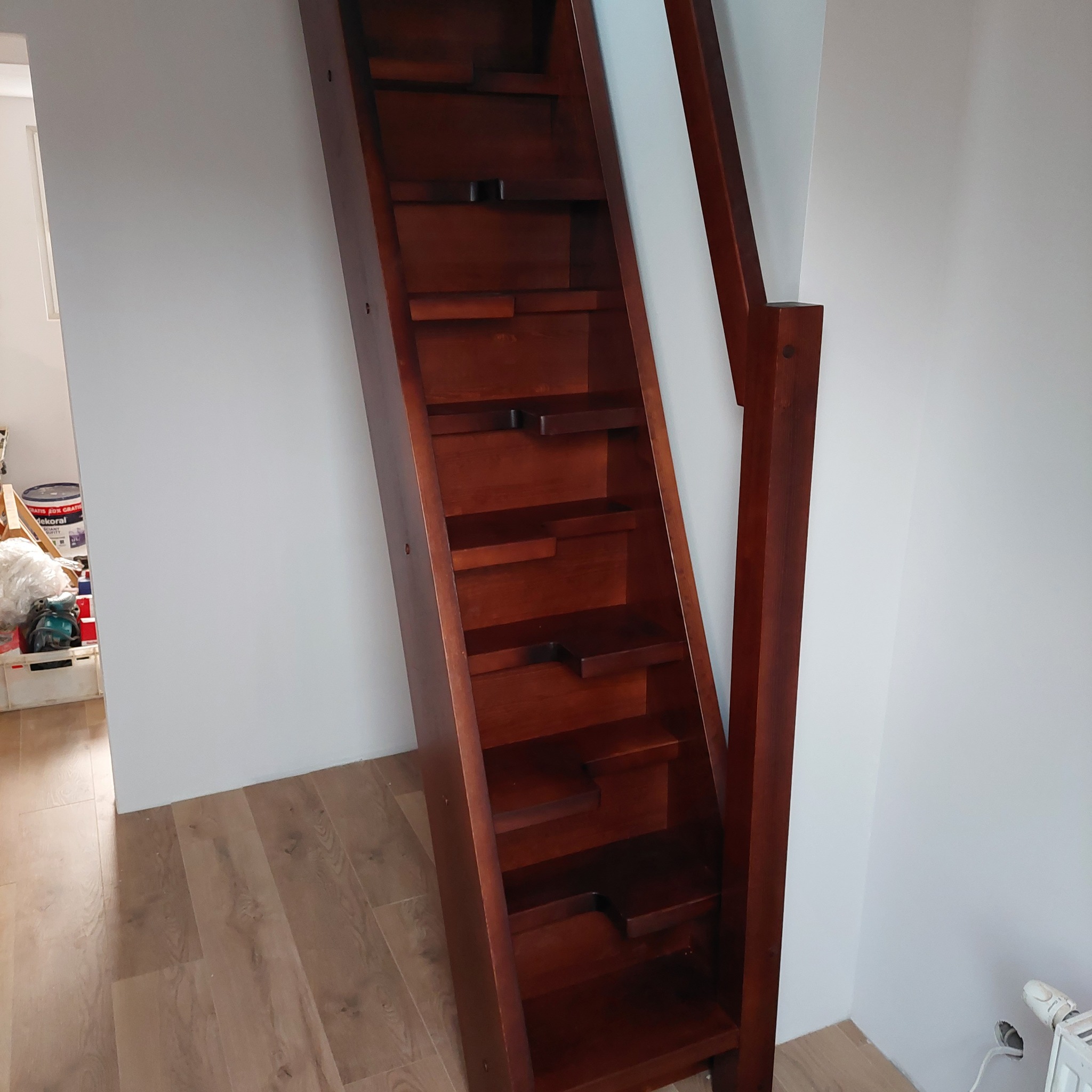 Kompaktowe, ciemnobrązowe schody o prostej konstrukcji, z widocznymi stopniami z wycięciami, zamontowane przy białej ścianie w jasnym wnętrzu.