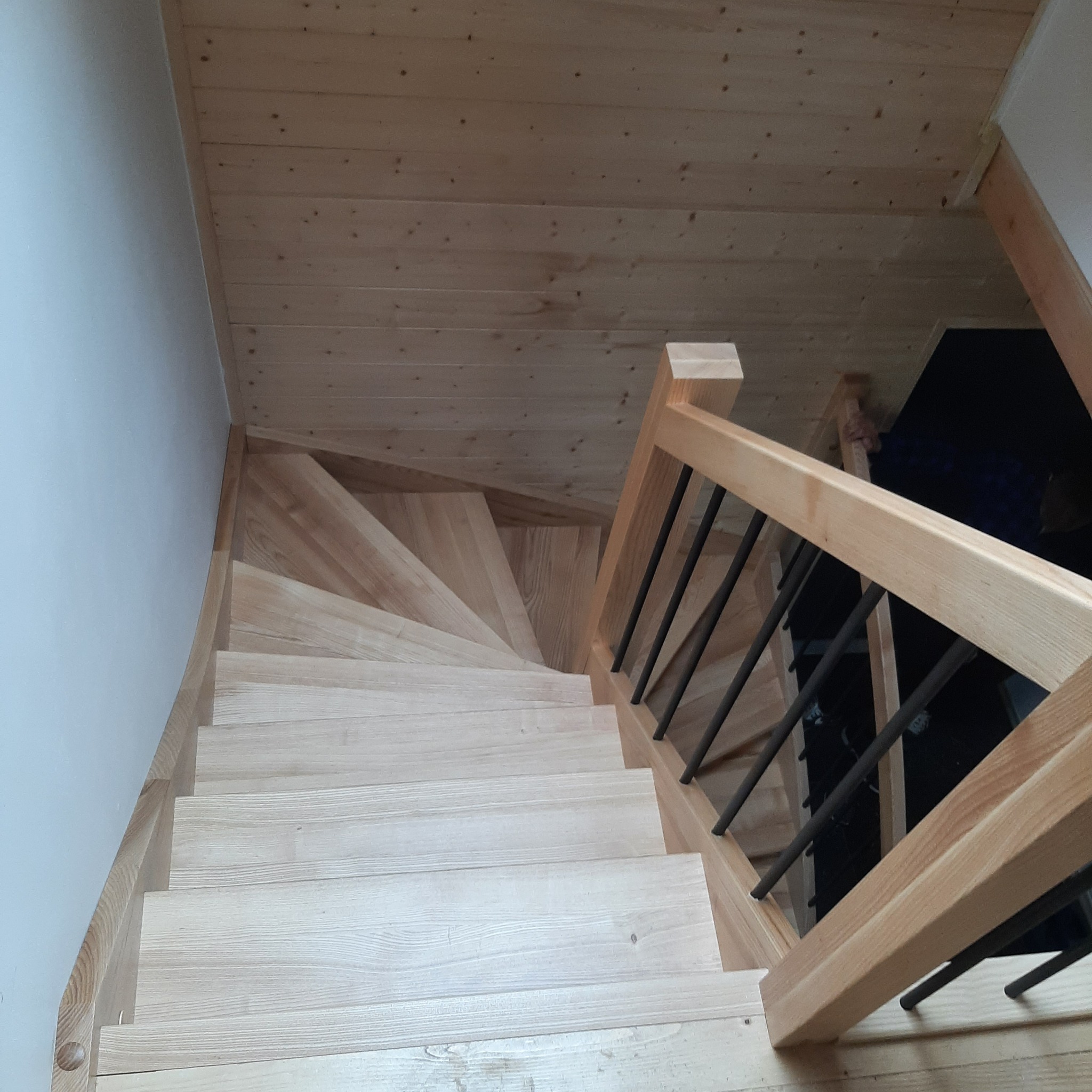 Jasne, spiralne schody z drewna z metalowymi prętami balustrady, widok z góry na drewnianą boazerię sufitową i białą ścianę.