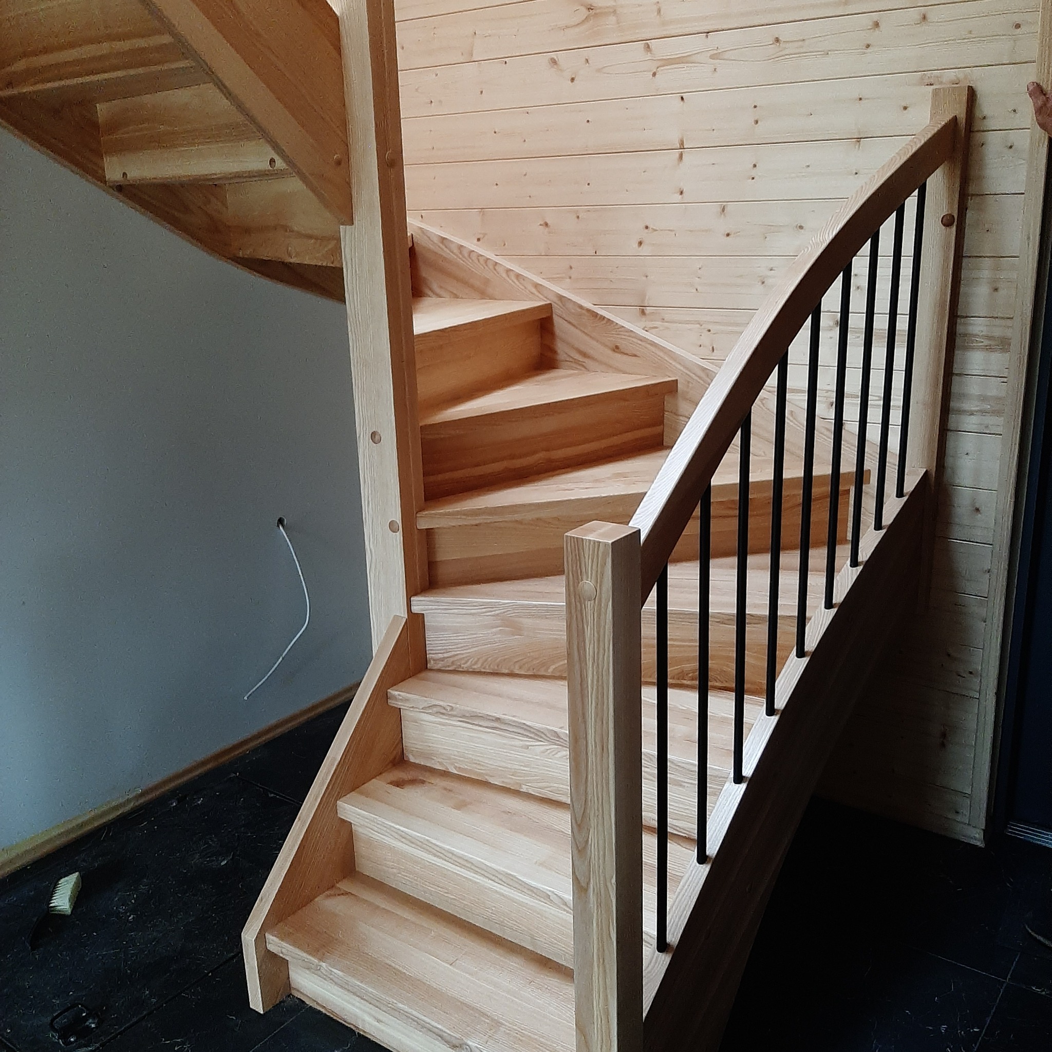 Jasne, drewniane schody z czarnymi, metalowymi balustradami, montowane przy ścianie wyłożonej drewnianą boazerią, widoczny kabel wystający ze ściany oraz szczotka na podłodze.