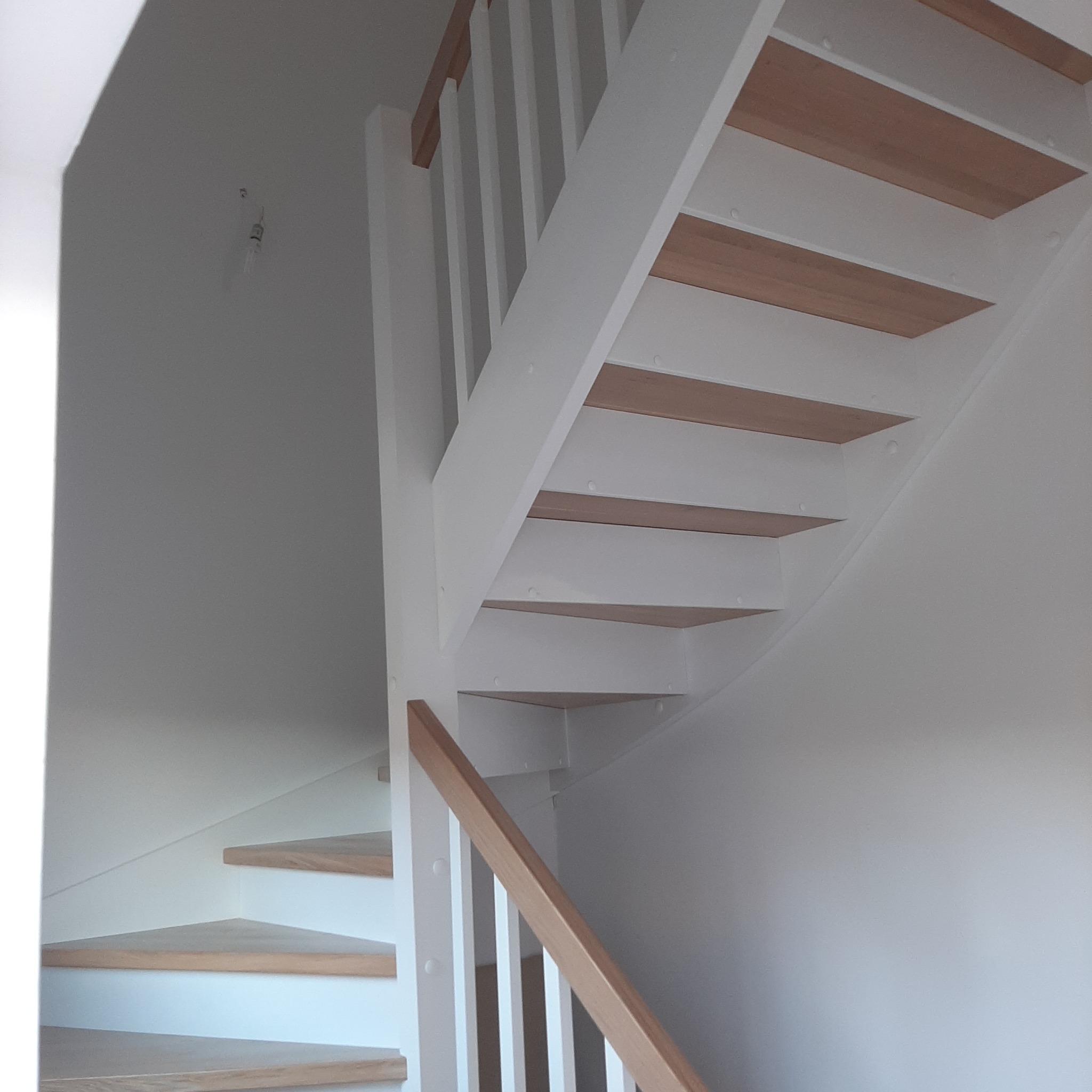 Białe schody z drewnianymi stopniami, widok od dołu, z minimalistyczną balustradą i surowym oświetleniem, w tle biała ściana z widocznym śladem po instalacji.