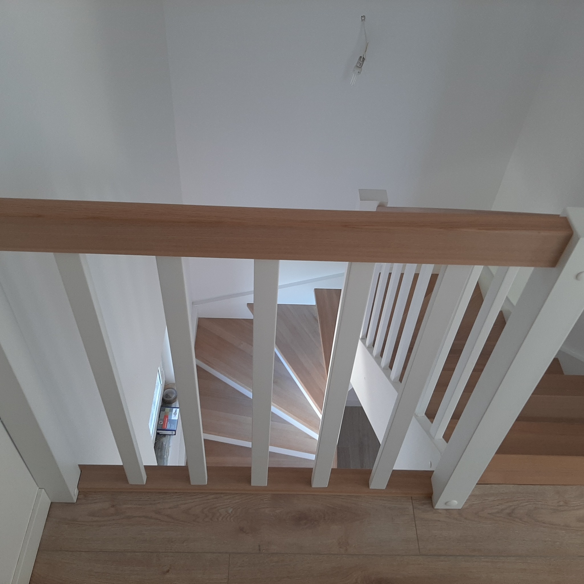 Widok z góry na spiralne schody z jasnego drewna z białymi balustradami, oświetlenie sufitowe widoczne w tle.