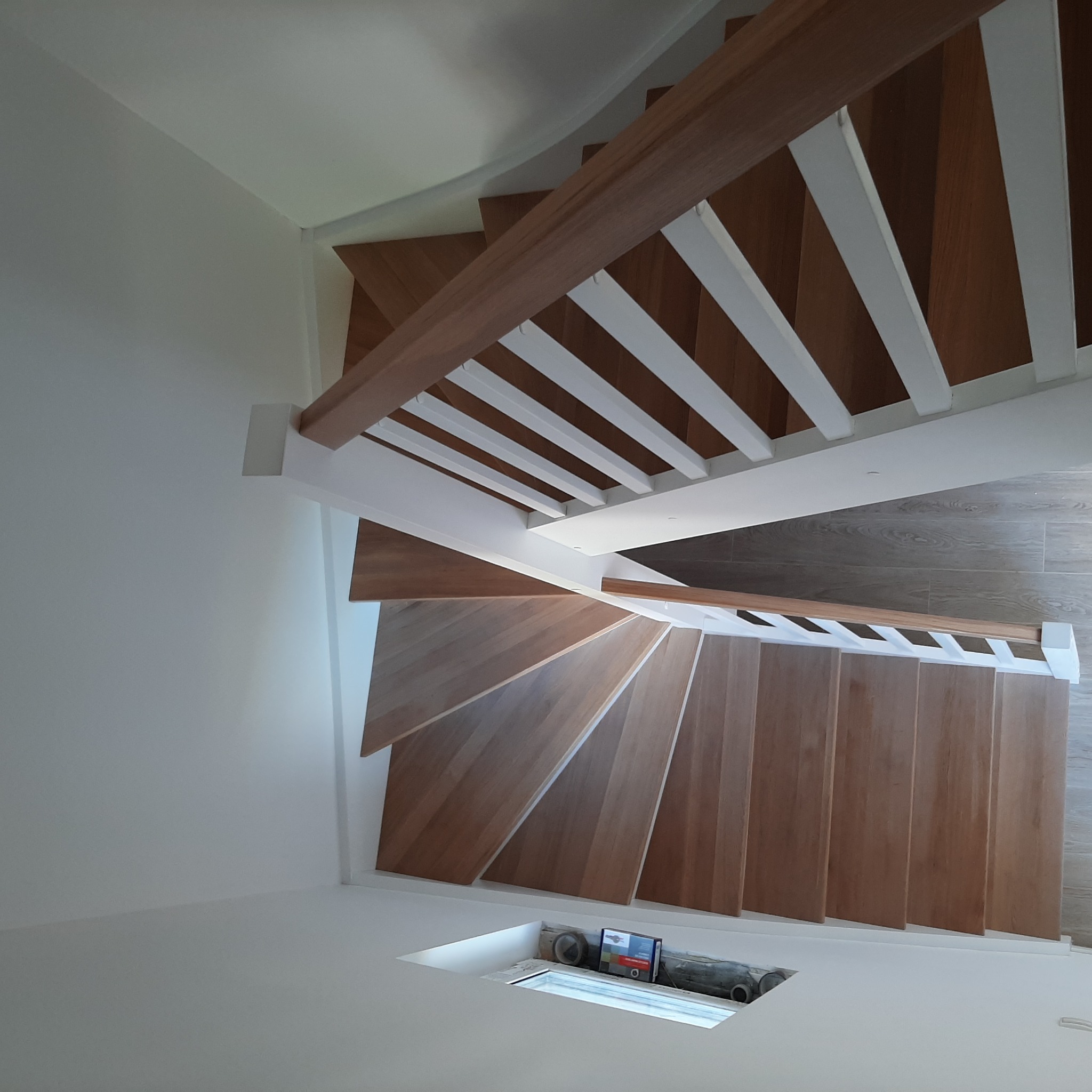 Spiralne schody z jasnego drewna z białymi balustradami, widok z góry, w tle płytki podłogowe i otwór w podłodze z widocznymi elementami instalacji.