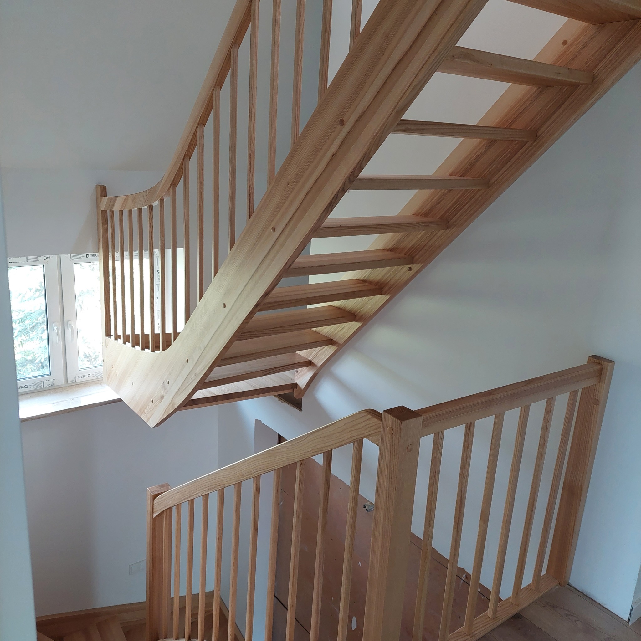 Jasne, drewniane schody z balustradą, widok z góry, naturalne oświetlenie, białe ściany, okno w tle.