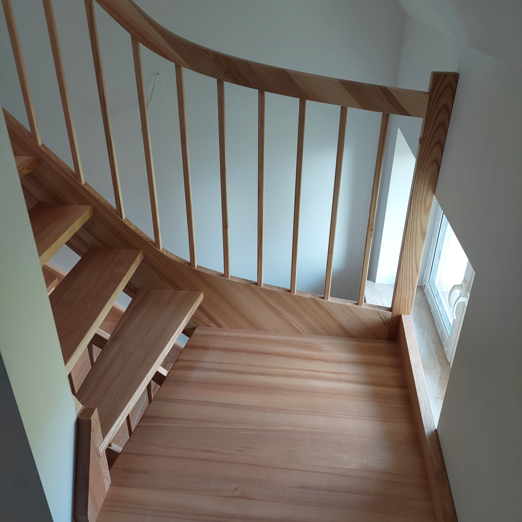 Jasne drewniane schody z giętą balustradą i pionowymi szczebelkami, widok z góry na dół, naturalne oświetlenie z okna obok.