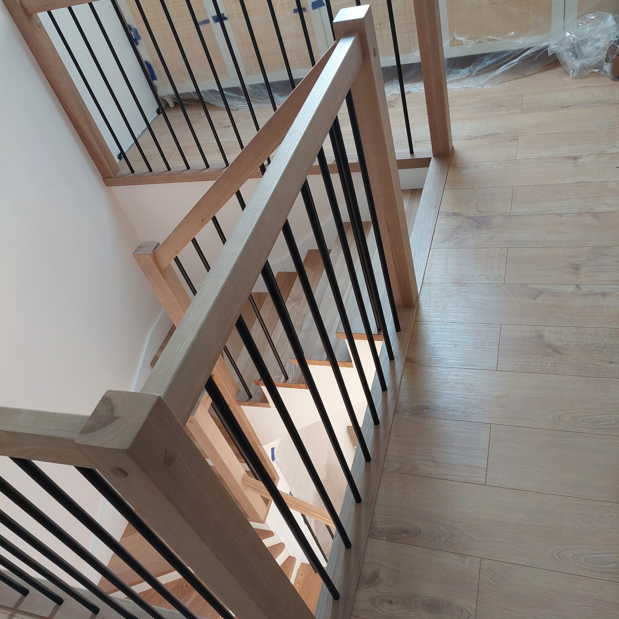 Nowoczesne schody z jasnego drewna z czarnymi, metalowymi tralkami, widok z góry na drewnianą podłogę i fragment zabezpieczonej folią ściany.