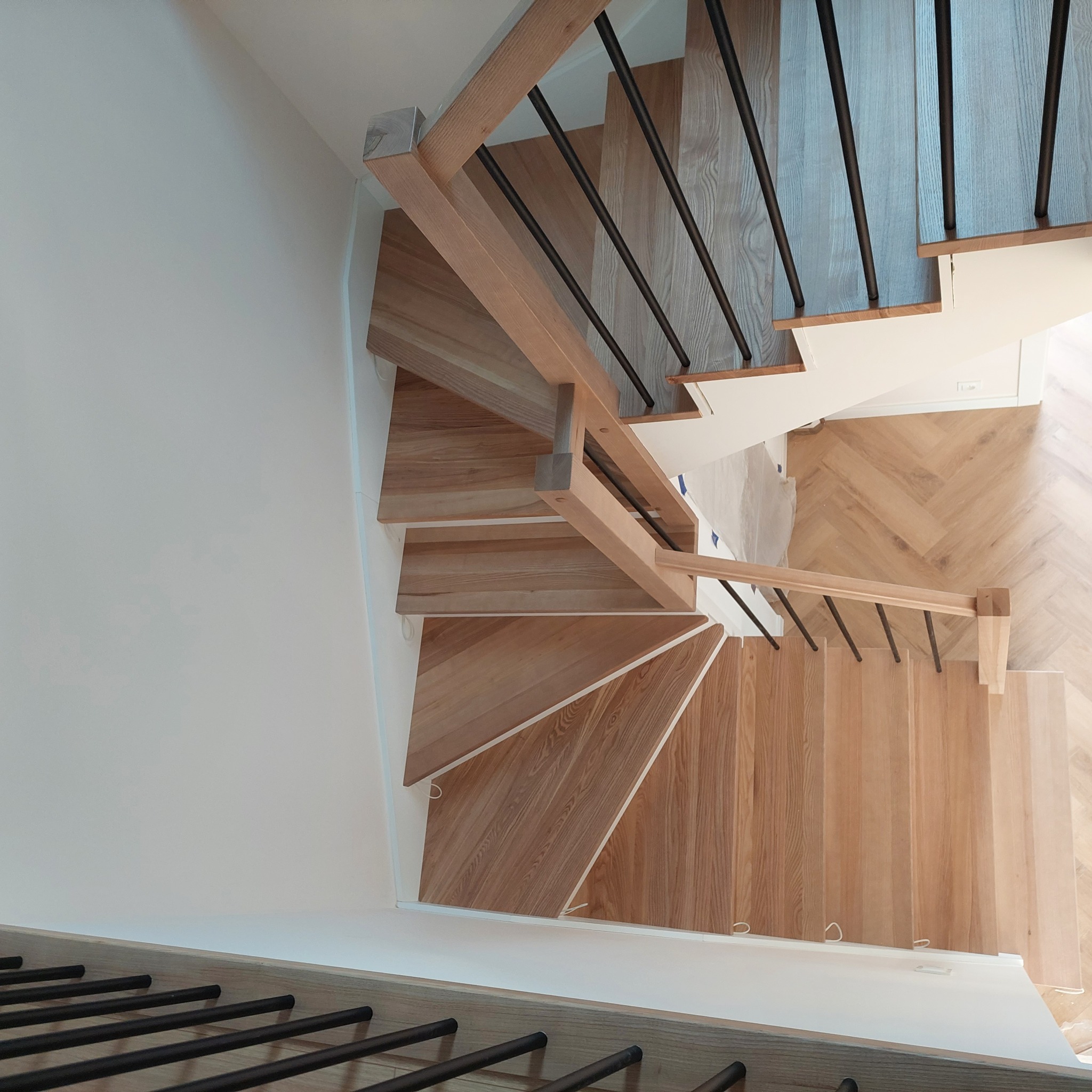 Widok z góry na jasne, drewniane schody z czarnymi, metalowymi poręczami, prowadzące w dół, z widoczną podłogą ułożoną w jodełkę.