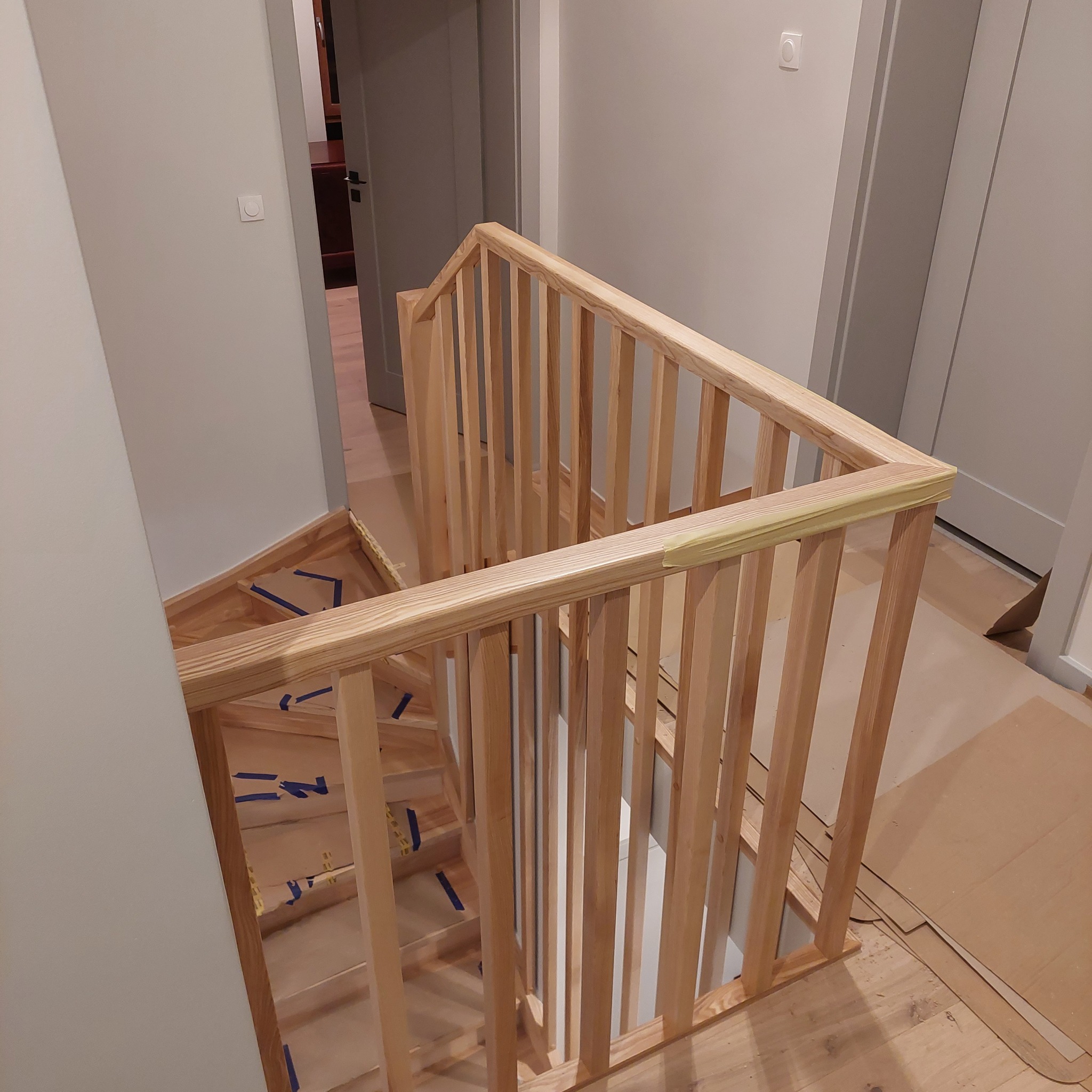 Surowe drewniane schody z balustradą zabezpieczoną taśmą malarską, widoczne zabezpieczenia podłogi kartonem, w tle szare ściany i drzwi.