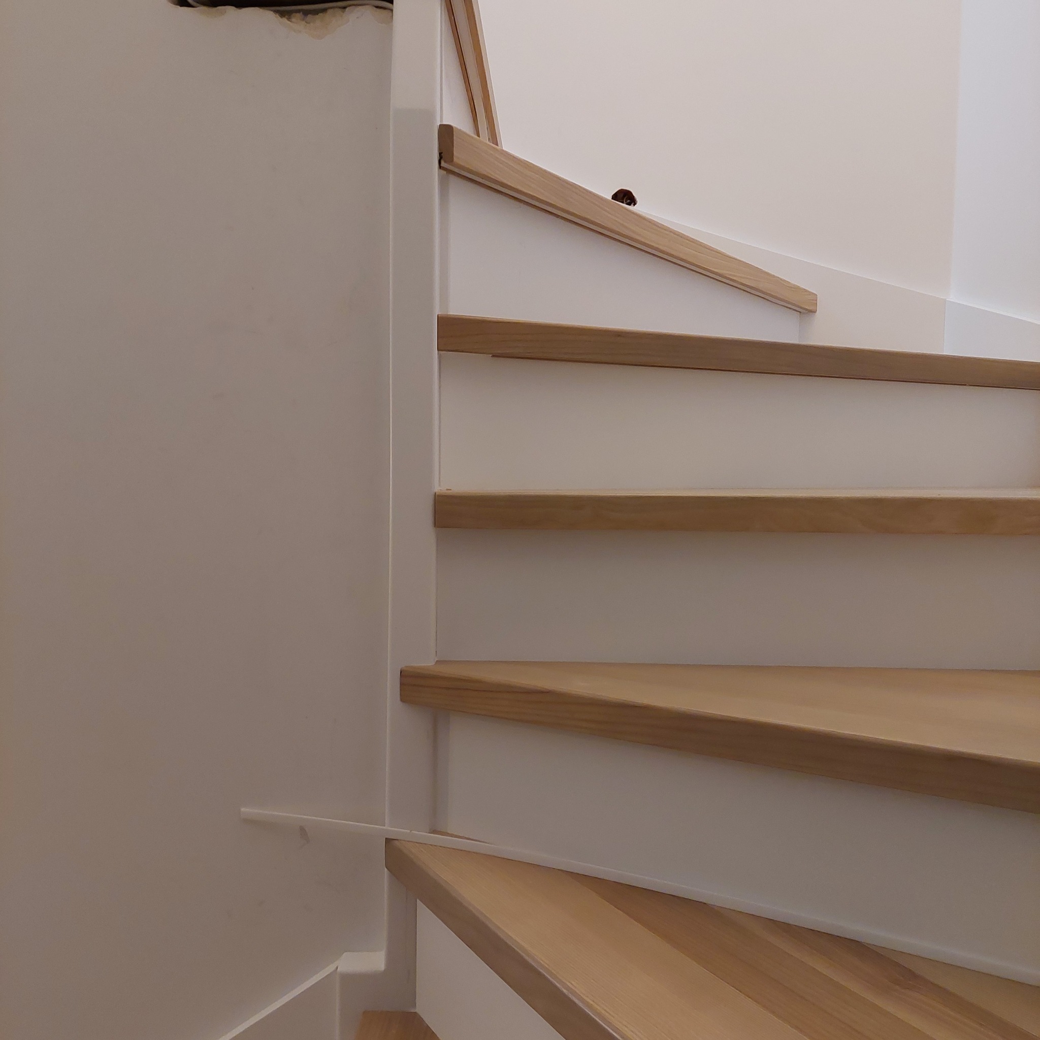 Jasne, drewniane stopnie schodów z białymi podstopnicami i listwą przypodłogową, widoczne niedociągnięcia w wykończeniu ściany na górze.