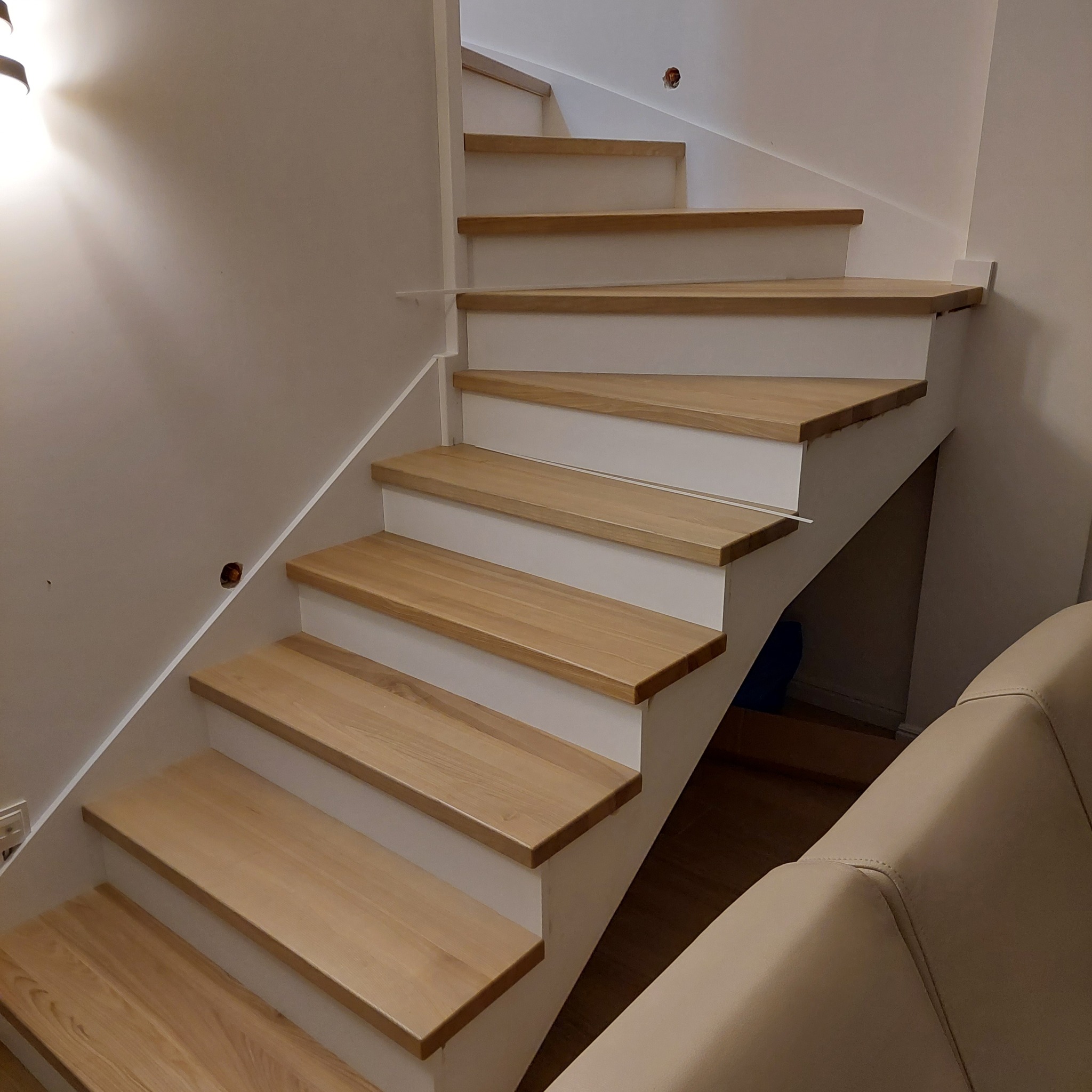 Jasne drewniane stopnie schodów z białymi podstopnicami, widoczne niedokończone otwory na ścianie oraz fragment beżowej sofy w tle.