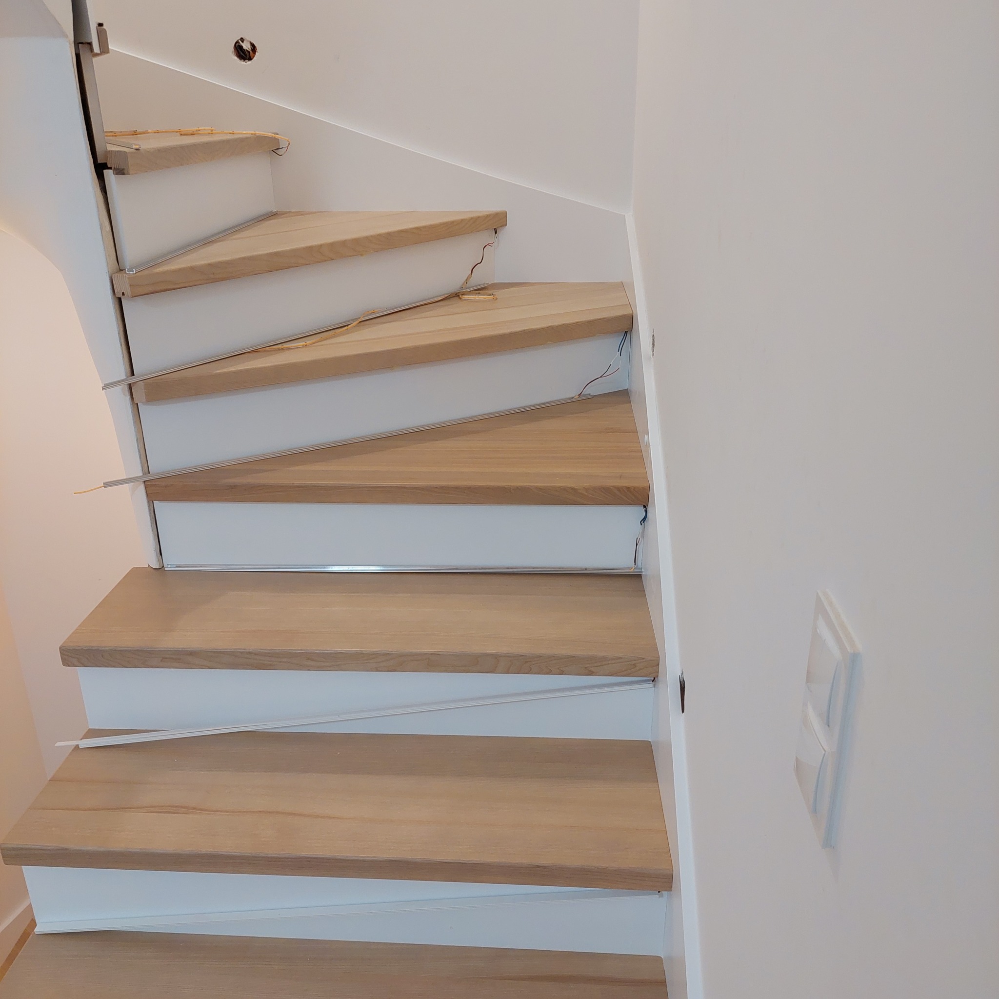 Bieg schodów z jasnego drewna z białymi podstopnicami, widoczne niedokończone podłączenia elektryczne i listwy oświetleniowe.