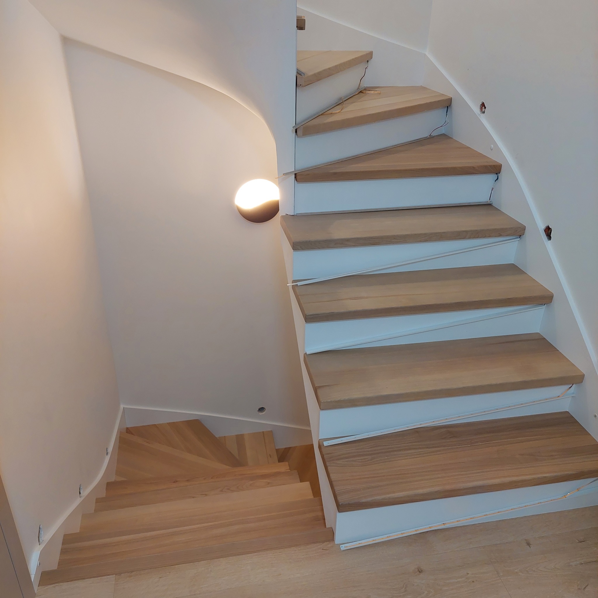 Drewniane schody spiralne z białymi podstopnicami i oświetleniem LED pod stopniami, widok z góry na dół.