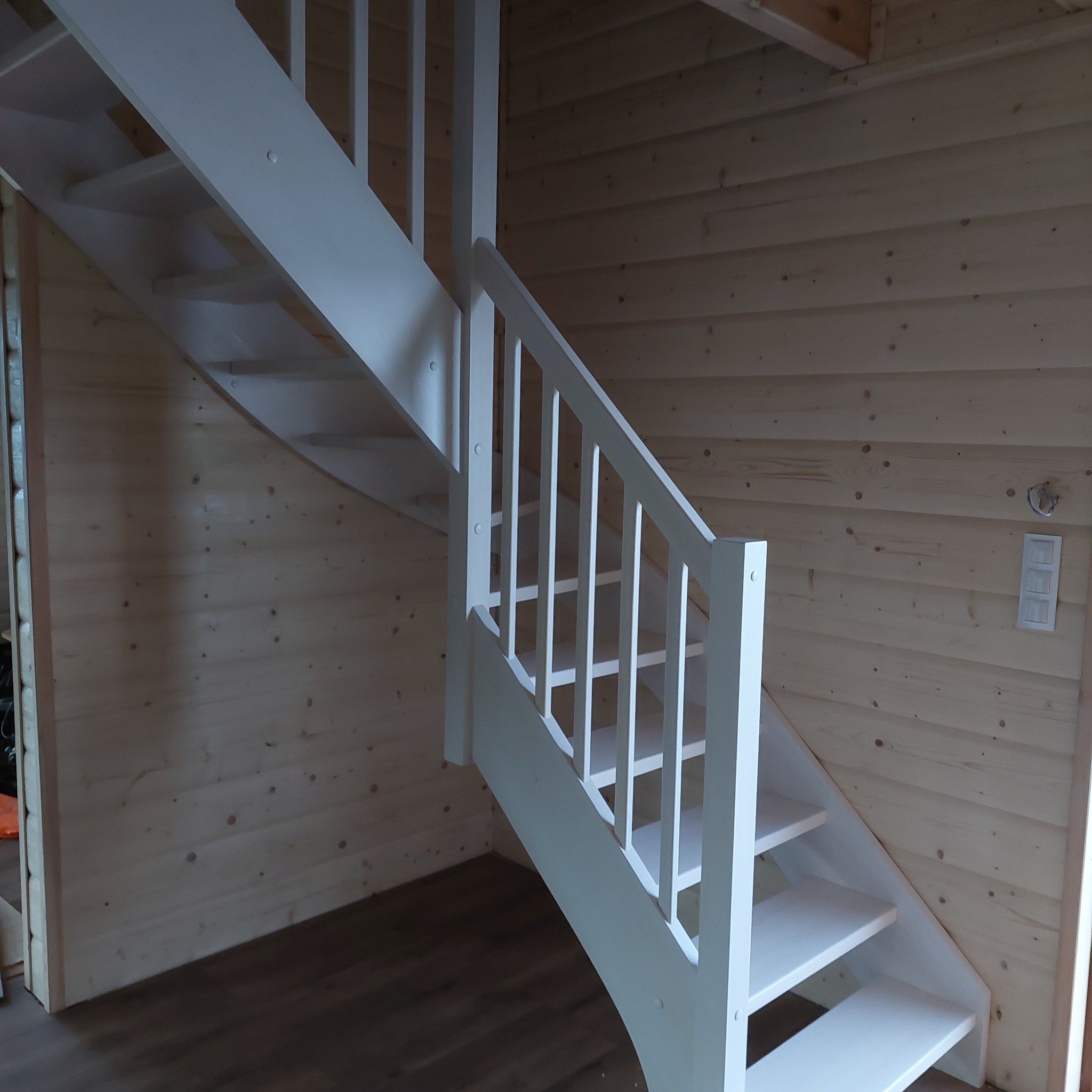 Białe schody z drewnianymi balustradami w domu z boazerią, widoczny włącznik światła na ścianie.