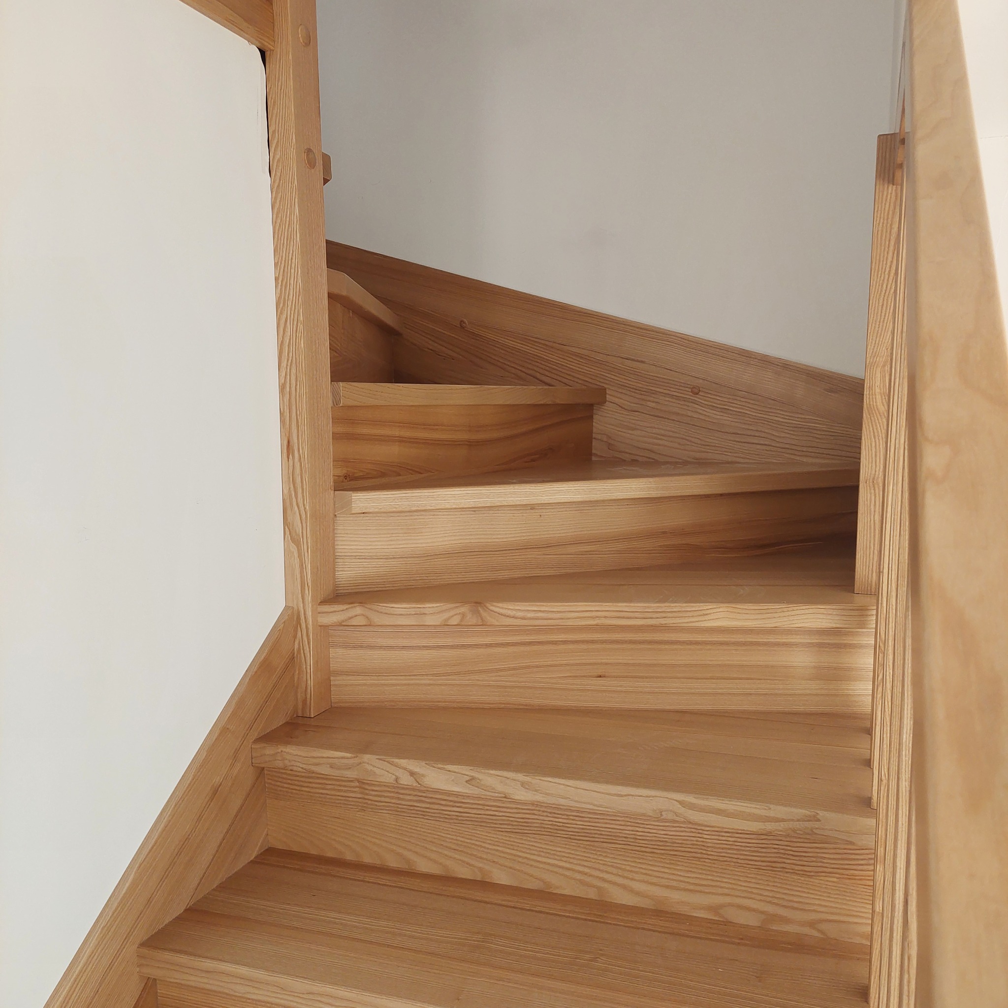 Jasne, lakierowane stopnie z drewna o wyraźnym usłojeniu, widok z dołu na bieg schodów przy białej ścianie.