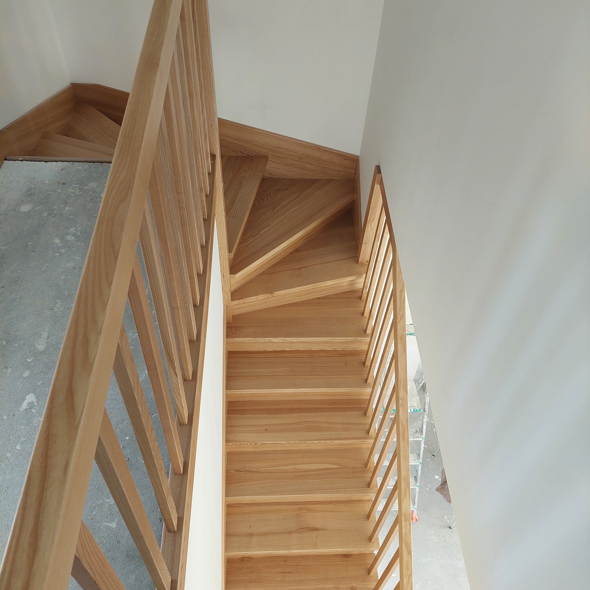 Jasne, dębowe schody z prostymi balustradami, widok z góry na zakręt schodów, surowy beton na podeście początkowym.