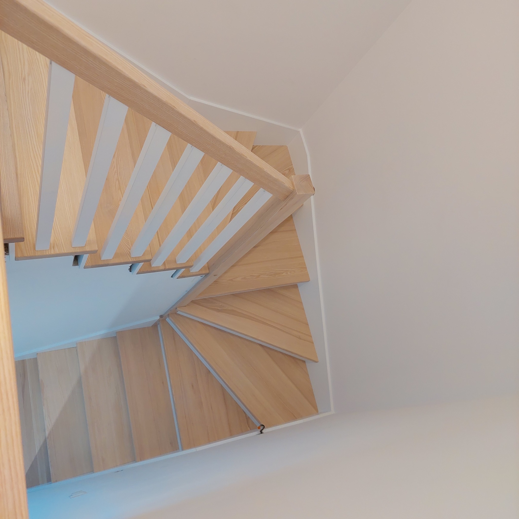 Widok z góry na drewniane schody spiralne z jasnego drewna z białymi balustradami w jasnym wnętrzu.