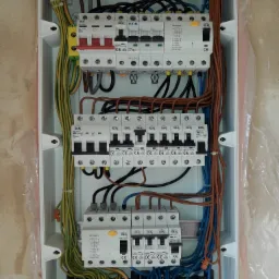 Otwarta skrzynka rozdzielcza z zamontowanymi bezpiecznikami i podłączonymi kablami w różnych kolorach, widoczne oznaczenia producentów i parametry techniczne urządzeń.