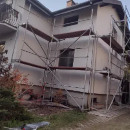 Dom w trakcie prac elewacyjnych, widoczne rusztowania, częściowo otynkowana fasada, okna zabezpieczone folią, materiały budowlane i narzędzia na trawniku.