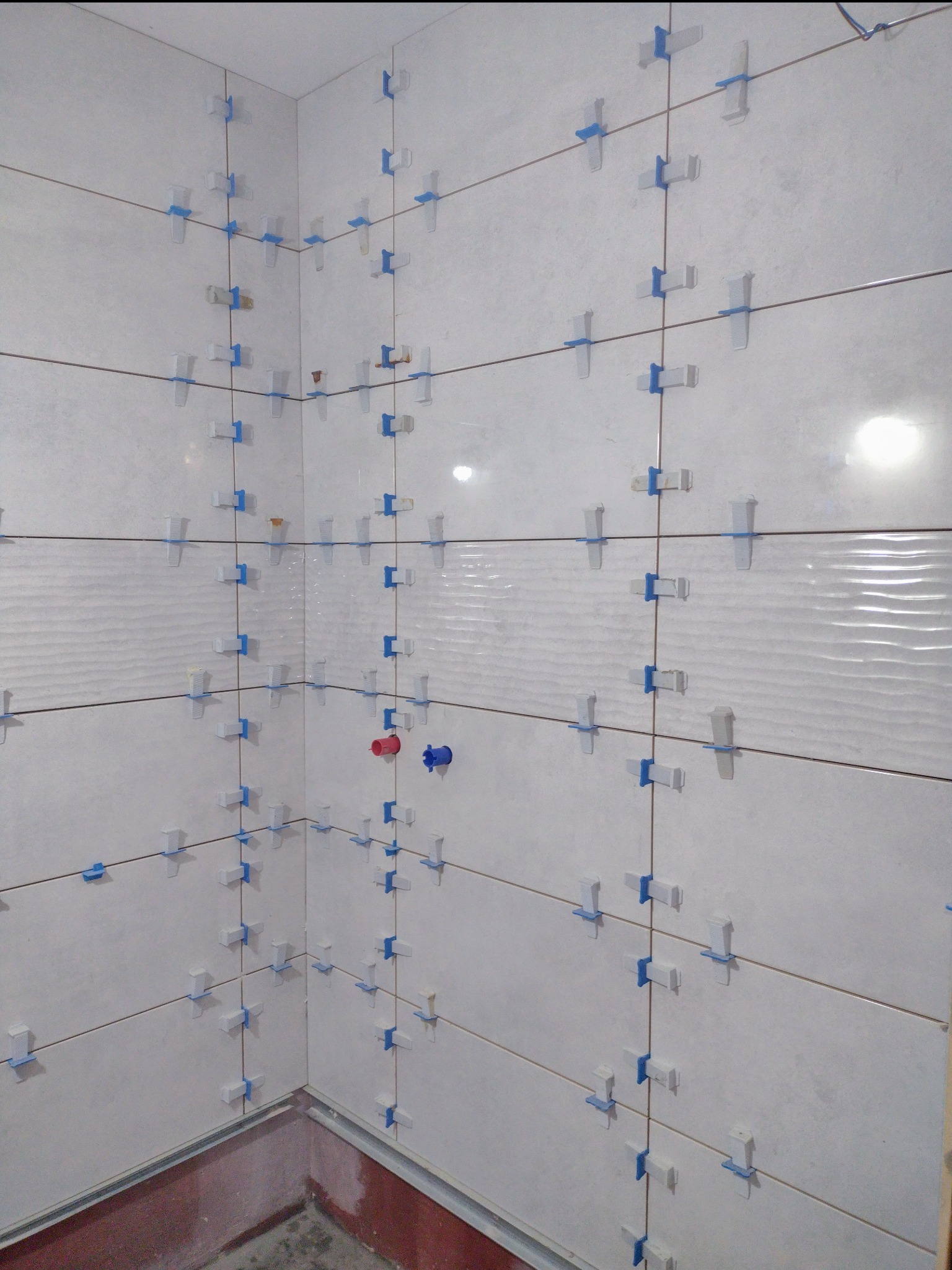 Świeżo ułożone białe płytki ceramiczne na ścianie w rogu pomieszczenia, z niebieskimi krzyżykami dystansowymi, widoczne przyłącza wodne.