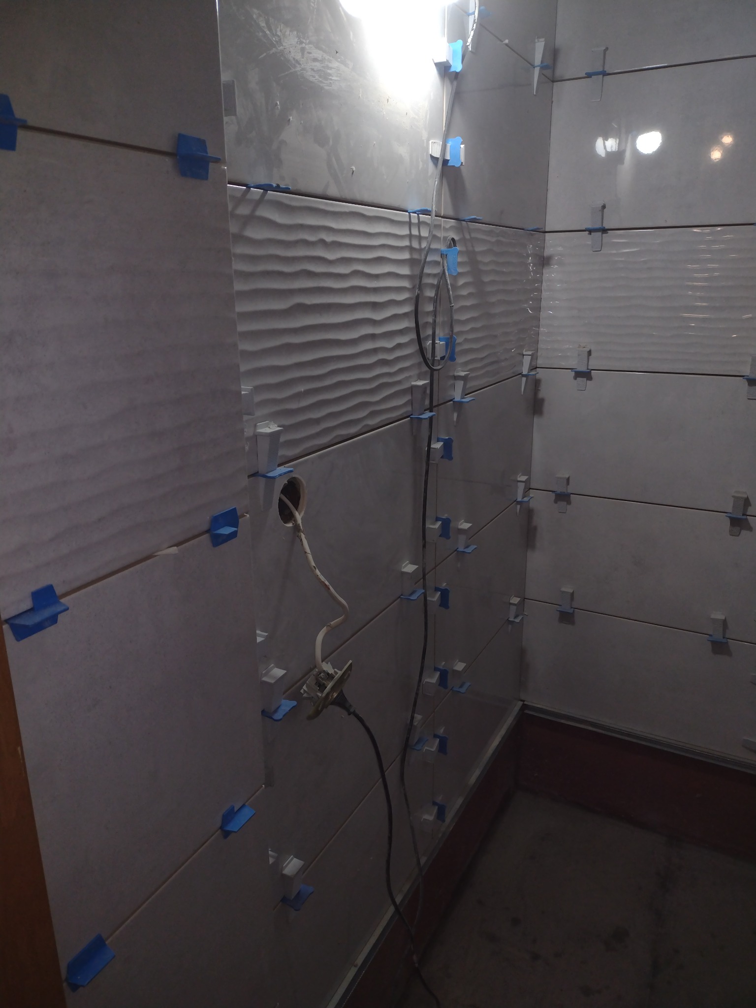 Świeżo ułożone, białe płytki ścienne z poziomym wzorem falistym, zabezpieczone niebieskimi krzyżykami dystansowymi, widoczny otwór na gniazdko elektryczne z wystającymi kablami i gniazdkiem,...