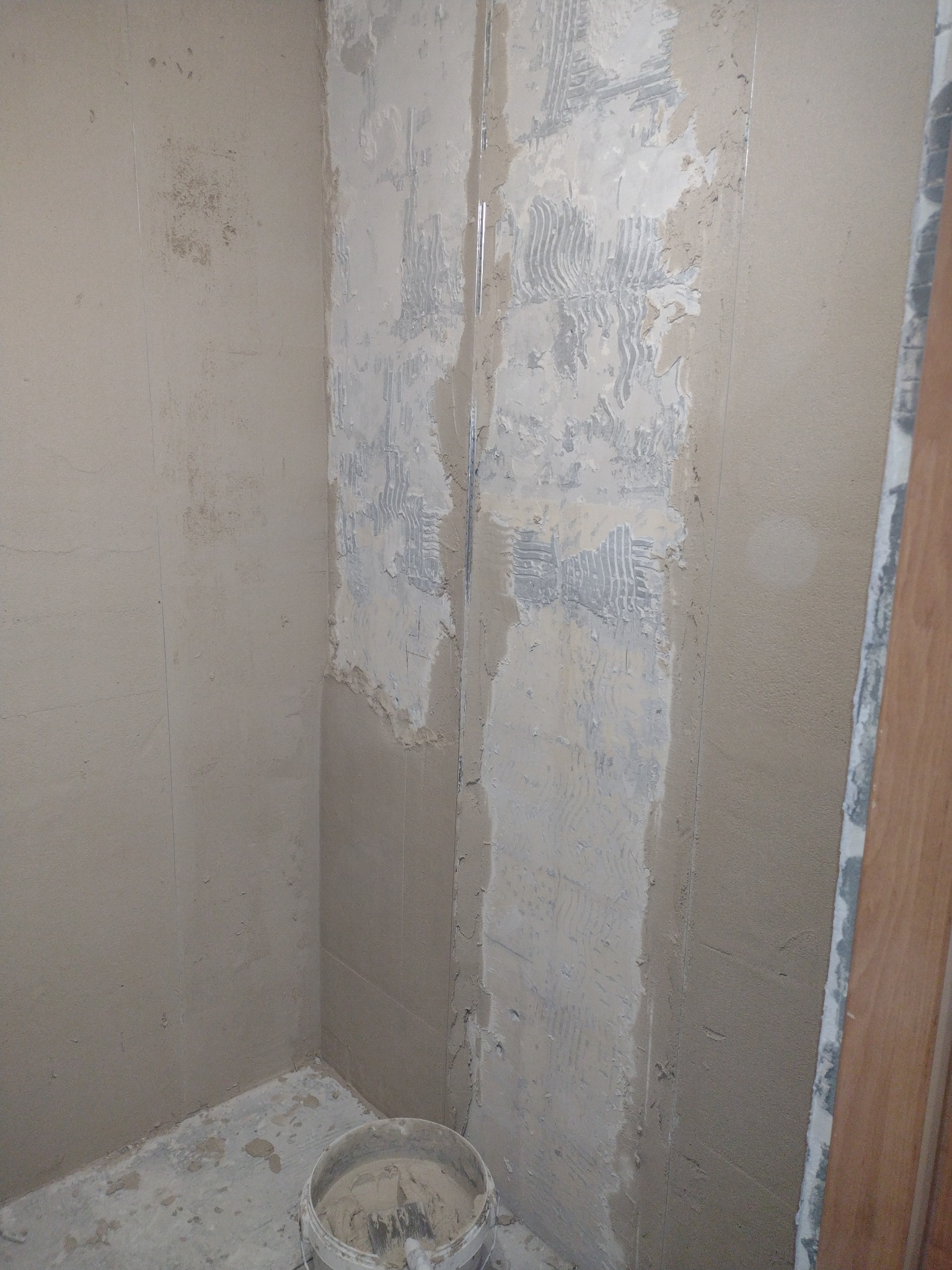 Przygotowanie ścian w łazience do położenia płytek, widoczny częściowo nałożony klej z charakterystycznymi śladami pacy oraz wiadro z zaprawą na podłodze.