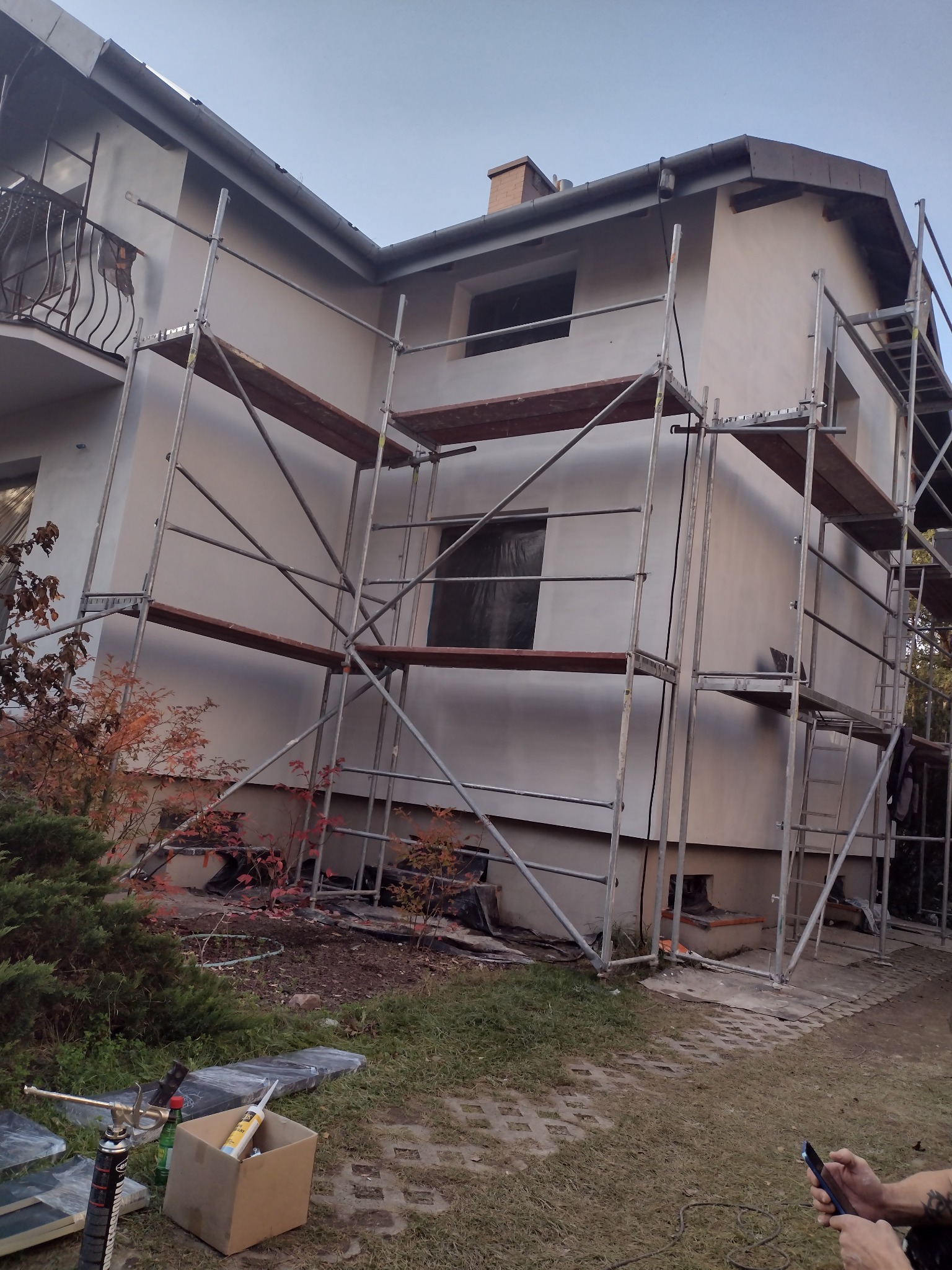 Dom w trakcie prac elewacyjnych, widoczne rusztowania, częściowo otynkowana fasada, okna zabezpieczone folią, materiały budowlane i narzędzia na trawniku.