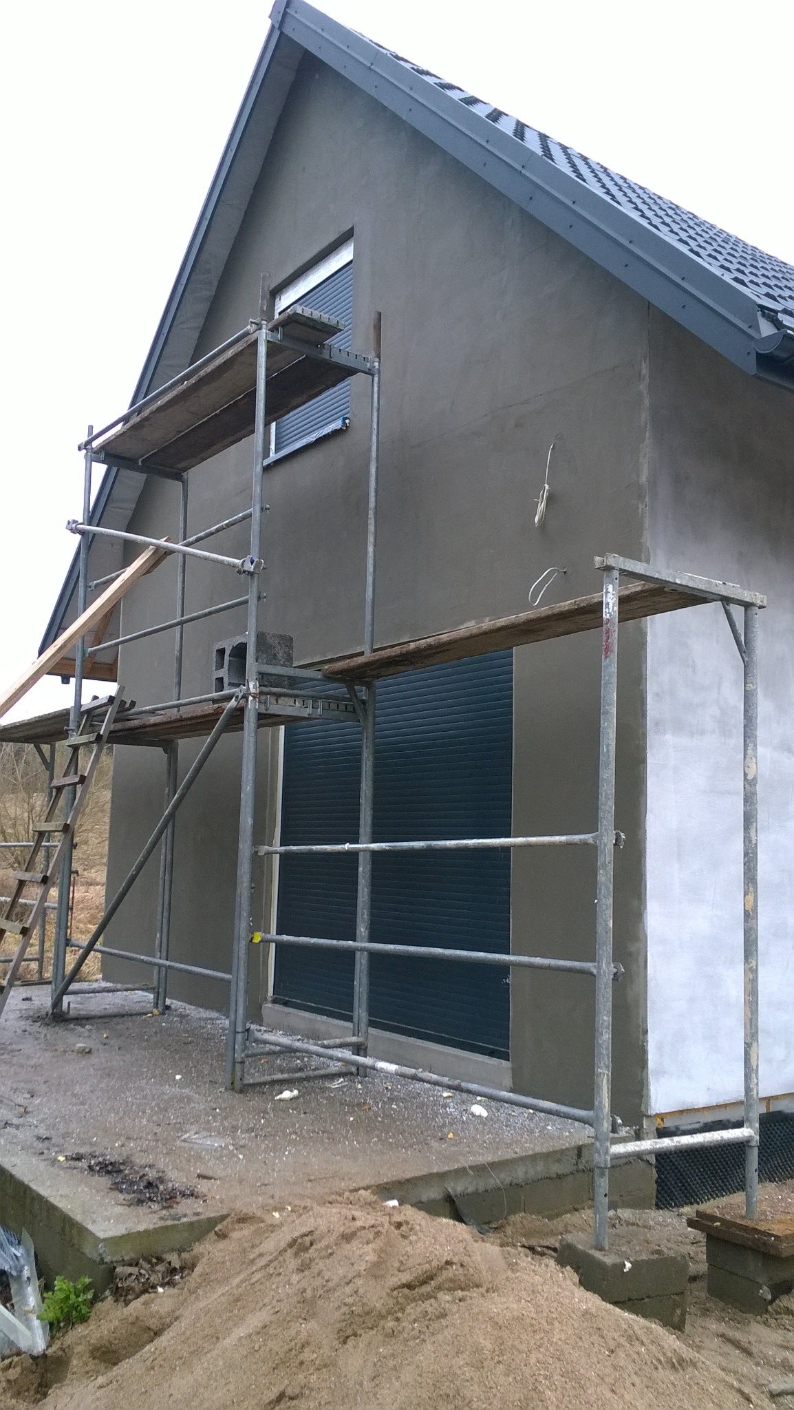 Dom w trakcie nakładania szarego tynku elewacyjnego, widoczne rusztowania i piasek na podłożu.