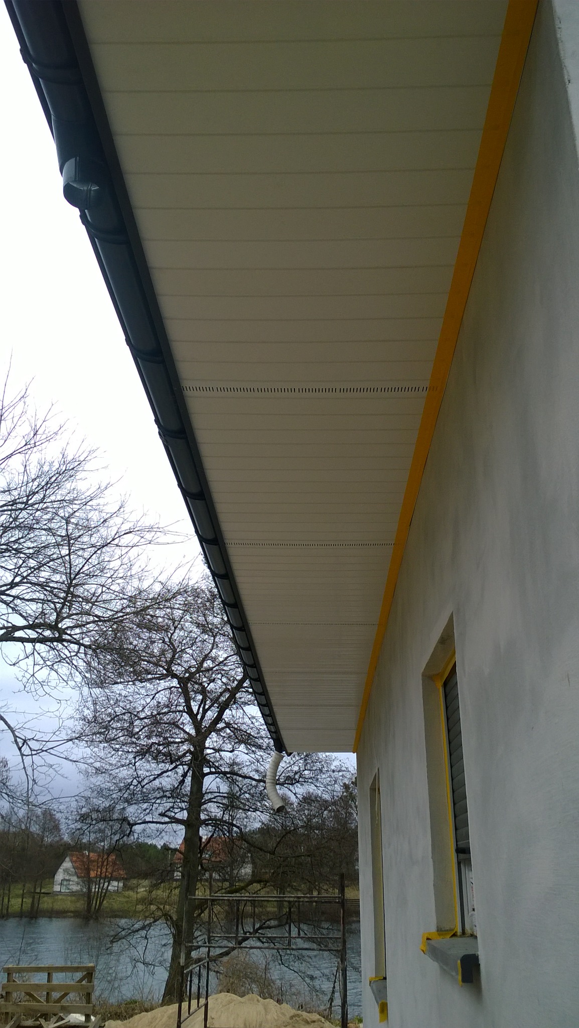 Wykończona podbitka dachowa z zamontowanym systemem rynnowym, widok z dołu na tle jeziora i drzew bez liści.