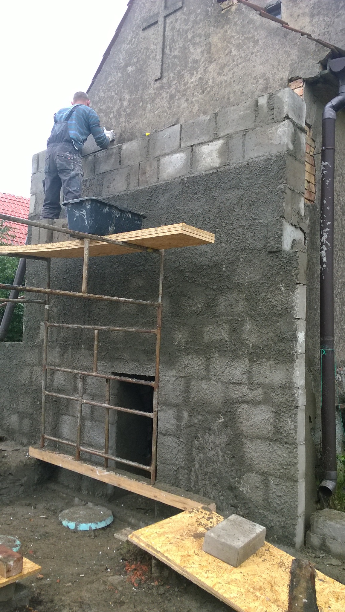 Pracownik na rusztowaniu, w trakcie murowania ściany budynku z betonu komórkowego, widoczny fragment elewacji z krzyżem.
