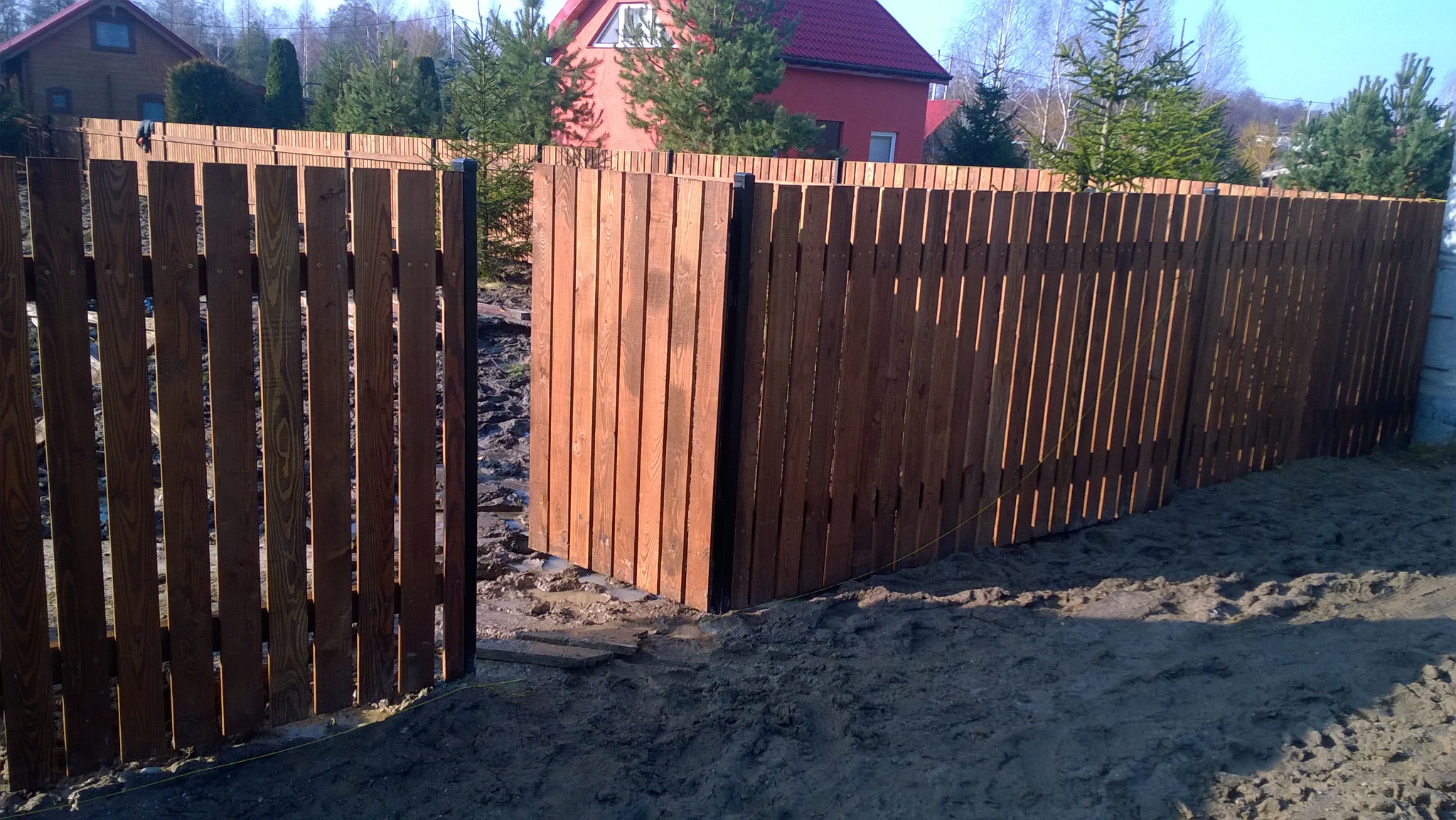 Nowo postawione drewniane ogrodzenie z pionowych desek, z czarnymi metalowymi słupkami, widoczne ślady prac ziemnych i dom w tle.