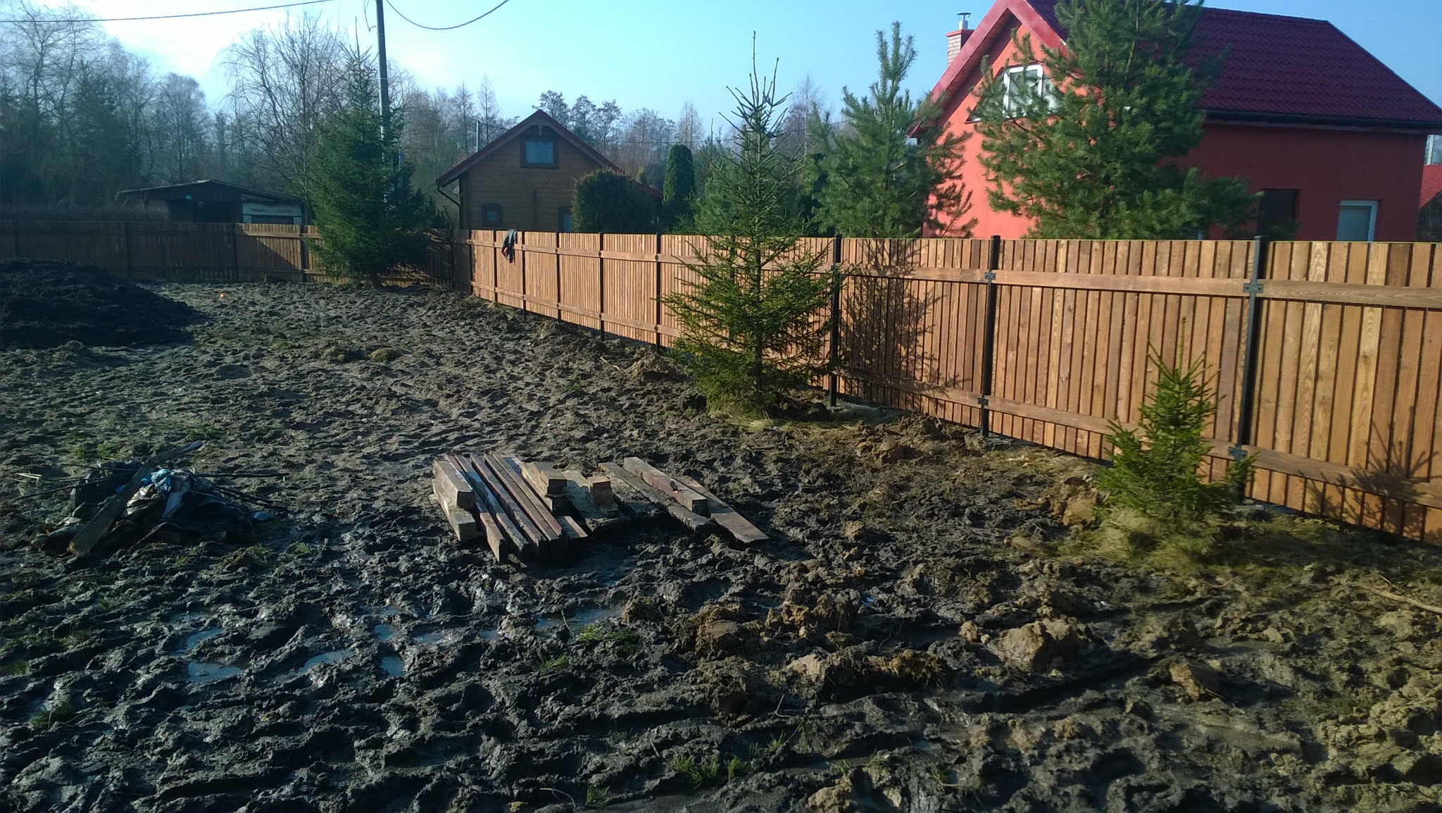 Nowy, drewniany płot panelowy oddziela posesję od sąsiadów, na pierwszym planie rozkopana, błotnista ziemia z pozostałościami materiałów budowlanych i młodymi drzewkami.