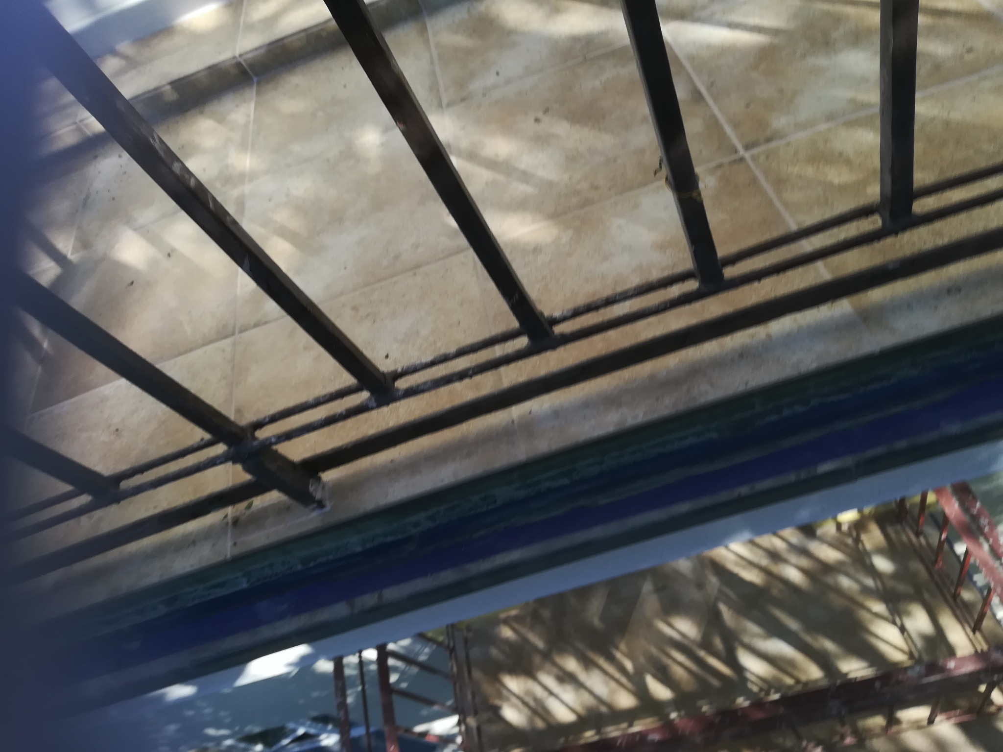 Metalowa konstrukcja zespawana z profili na tle beżowych płytek podłogowych, widok z dołu, częściowo zasłonięta przez krawędź balkonu.