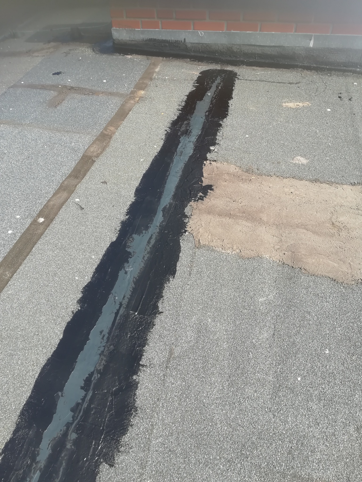 Uszczelnianie dylatacji na płaskim dachu pokrytym papą, widoczne świeże, czarne wypełnienie i fragment muru w tle.