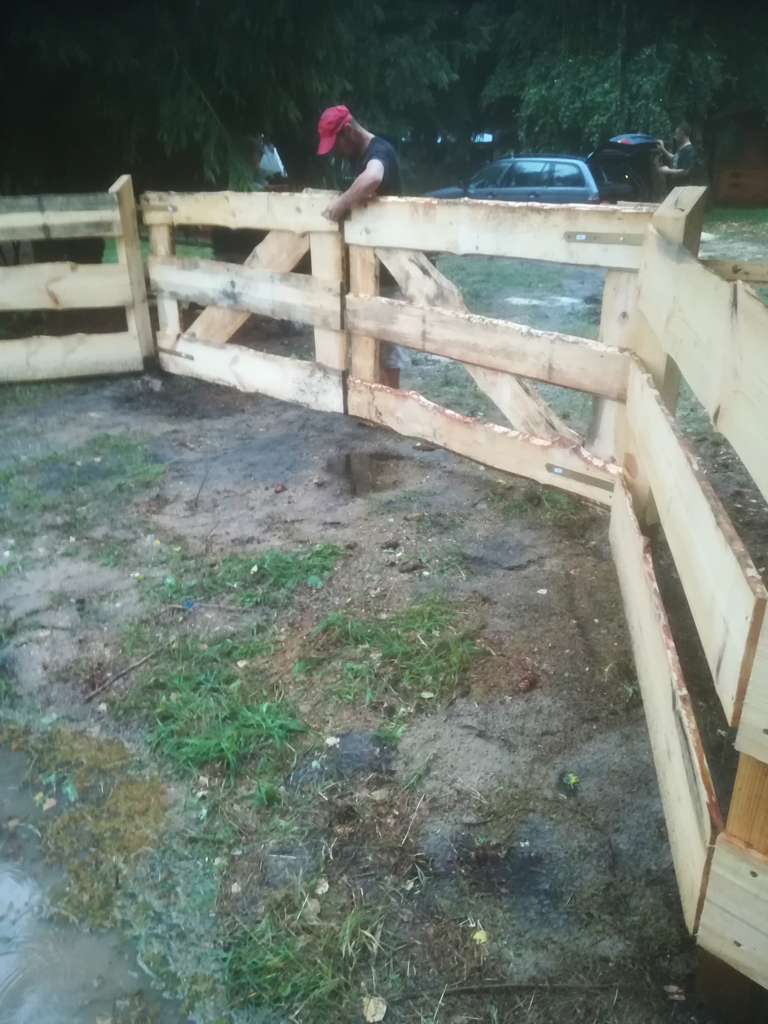 Mężczyzna w czerwonej czapce montuje drewnianą bramę z surowych desek, widoczny teren z błotem i trawą, w tle zaparkowany samochód.