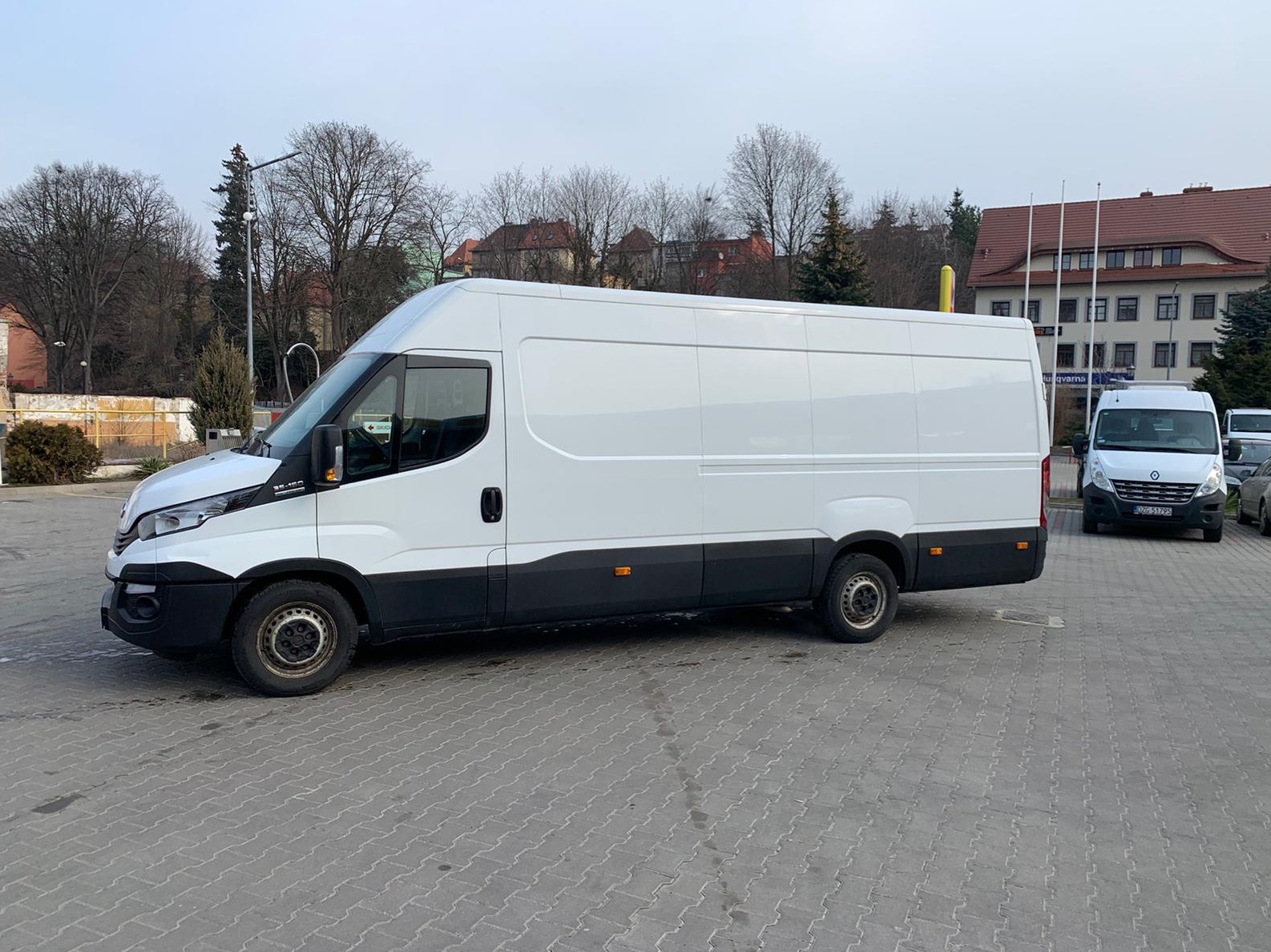 Biały furgon Iveco z czarnym dołem zaparkowany na kostce brukowej, z drzewami bez liści i budynkiem w tle, w pochmurny dzień.