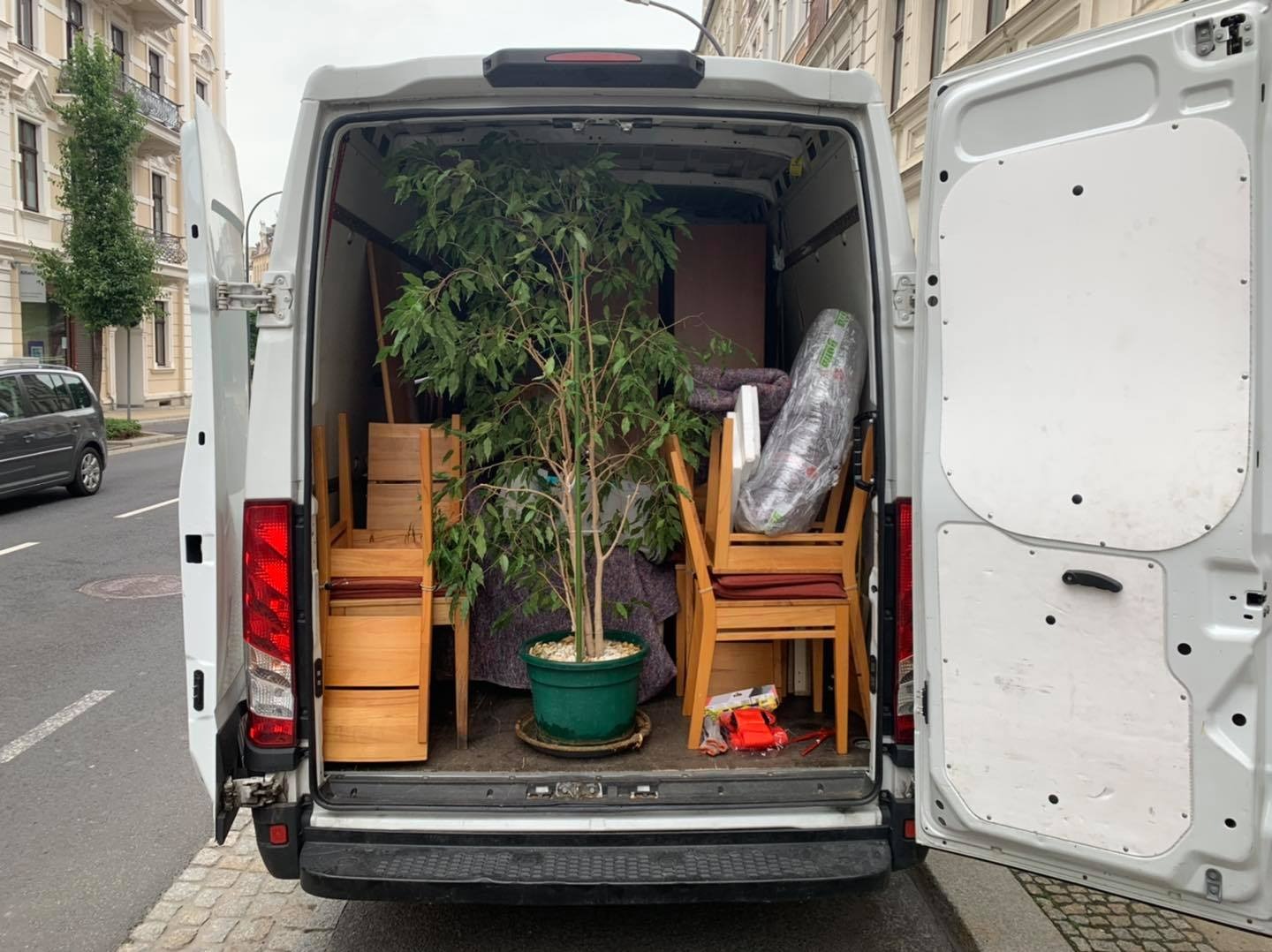 Wnętrze białego busa załadowane meblami, krzesłami z czerwonymi poduszkami, zapakowanym materacem i dużym kwiatem doniczkowym w zielonej donicy podczas transportu po ulicy miasta.