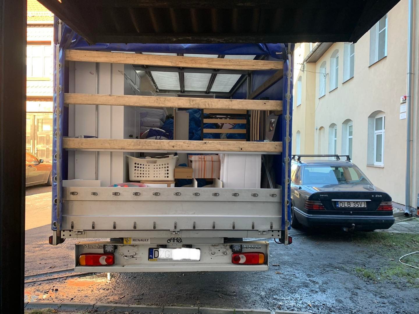 Tył otwartego samochodu dostawczego Renault, wypełnionego meblami i kartonami, zaparkowanego na zewnątrz budynku z jasną elewacją; obok stoi czarny Mercedes E 250 z bagażnikiem dachowym.