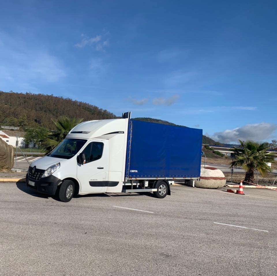 Biały bus z niebieską plandeką zaparkowany na parkingu, w tle góry i palmy.
