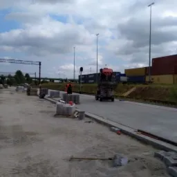 Trwa układanie nawierzchni z kostki brukowej na terenie przemysłowym, widoczne palety z kostką, maszyna do transportu, pracownicy w odblaskowych kamizelkach i kontenery w tle.