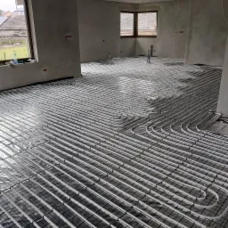 Rozległa instalacja ogrzewania podłogowego z białych rurek na srebrnej folii refleksyjnej w surowym wnętrzu z oknami i wystającymi rurami.